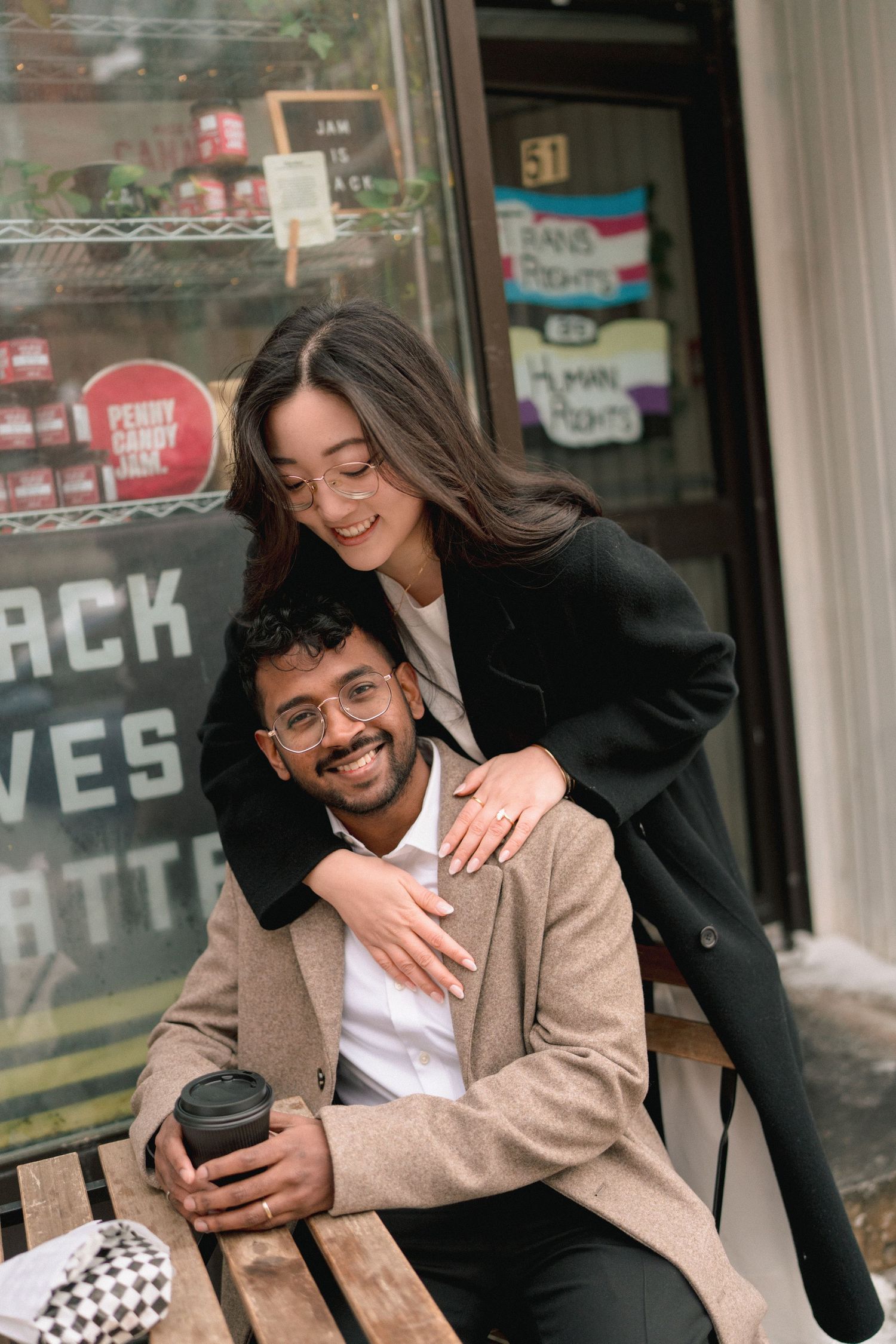 A couple in black and beige outfits sharing a playful moment while looking at a phone outdoors.