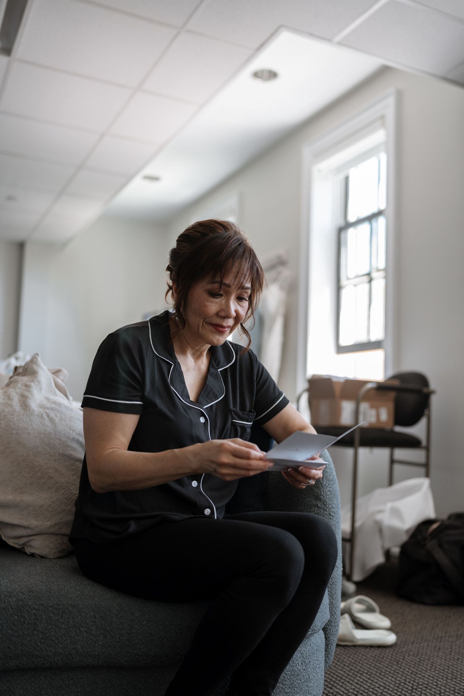 A person in black silk pajamas reads a document while sitting on a gray couch in a bright room.