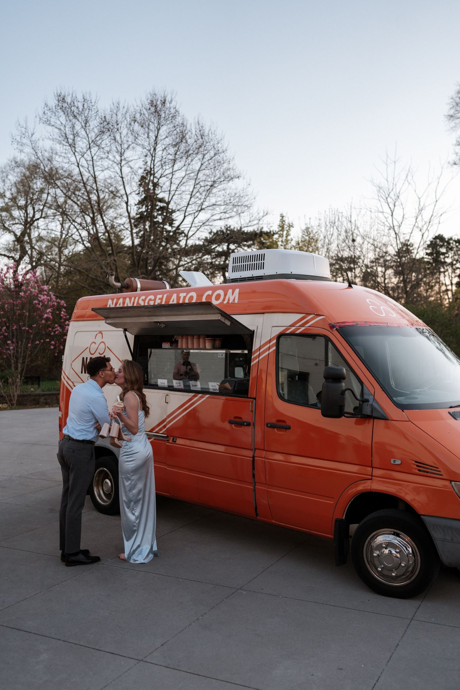 A bright orange food truck parked outdoors serving customers in formal attire at dusk.