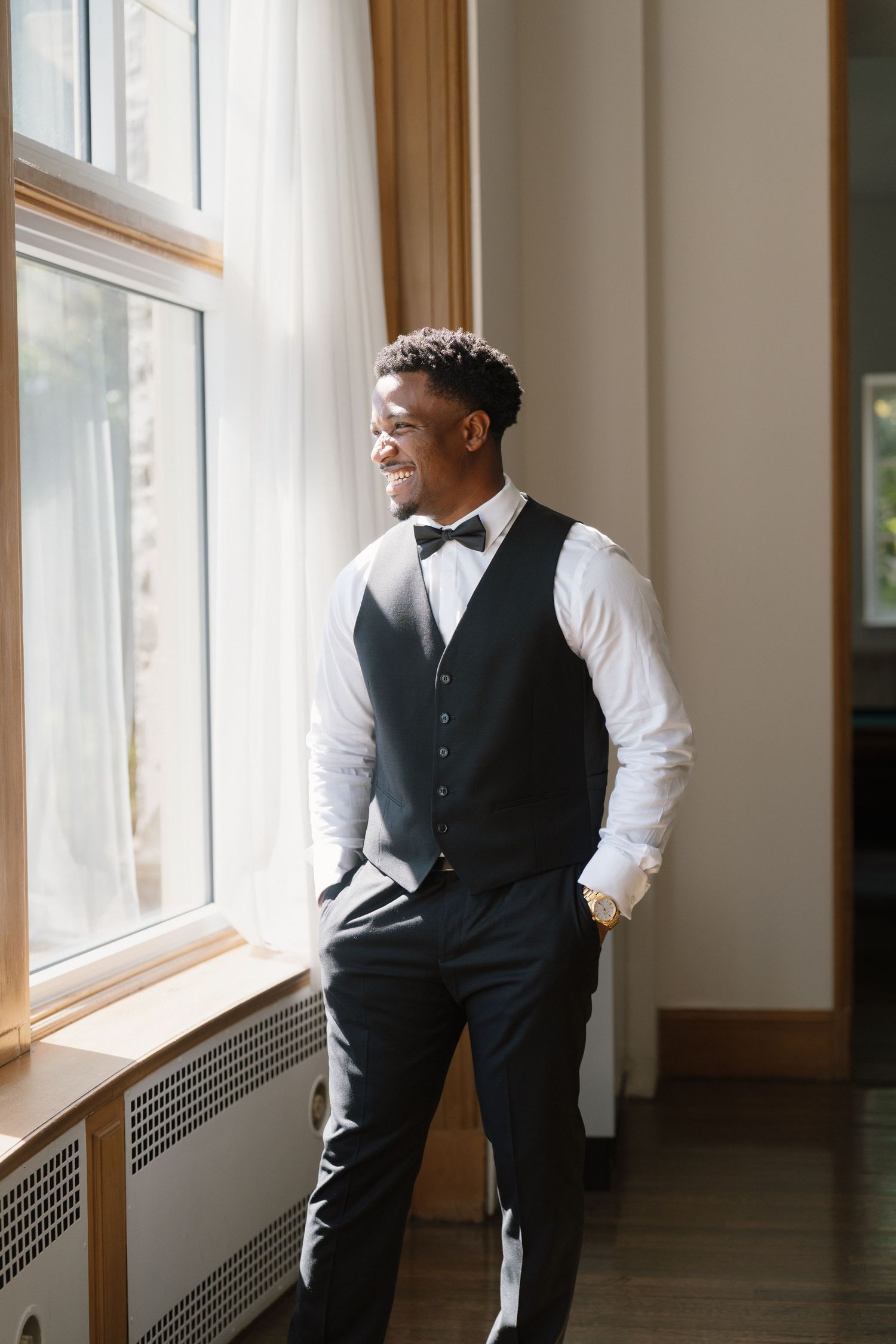 Smiling groom in black vest, bow tie, and white shirt stands by sunlit window with hands in pockets.