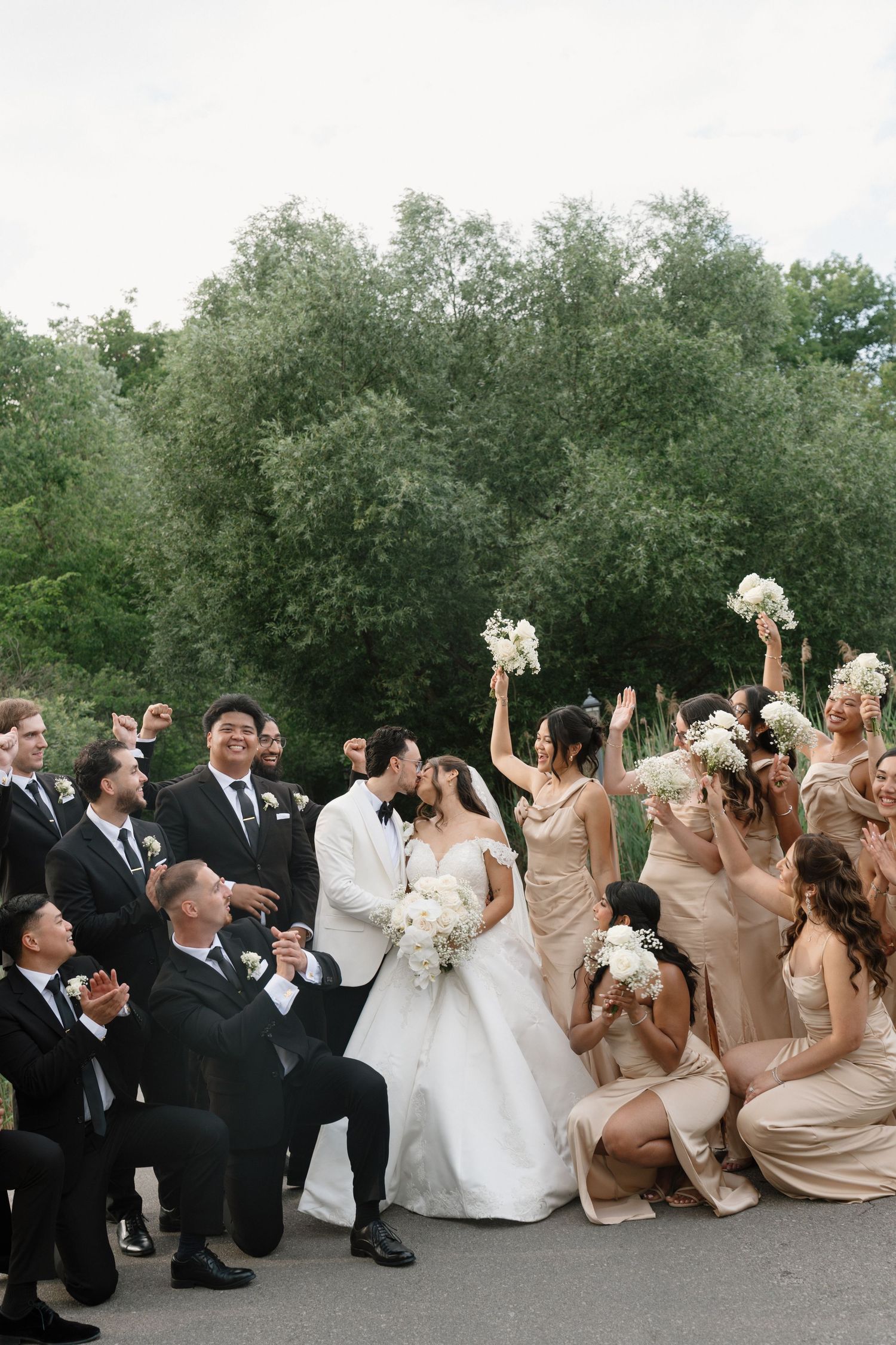 Wedding party celebrates as bride and groom kiss, bridesmaids in champagne gowns holding white bouquets, groomsmen in black suits.