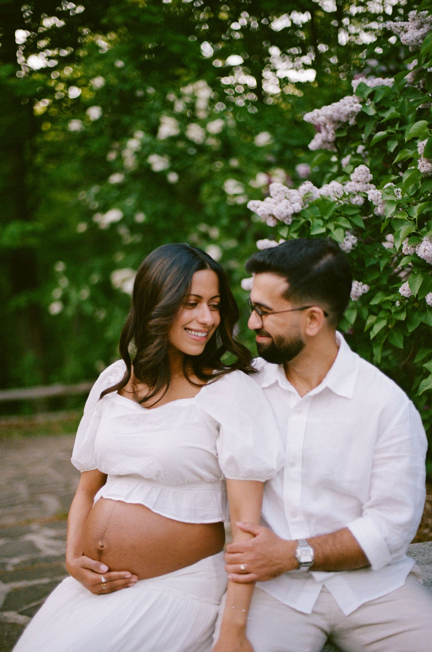 Expecting couple in white outfits smiling together outdoors near blooming lilac bushes in spring.