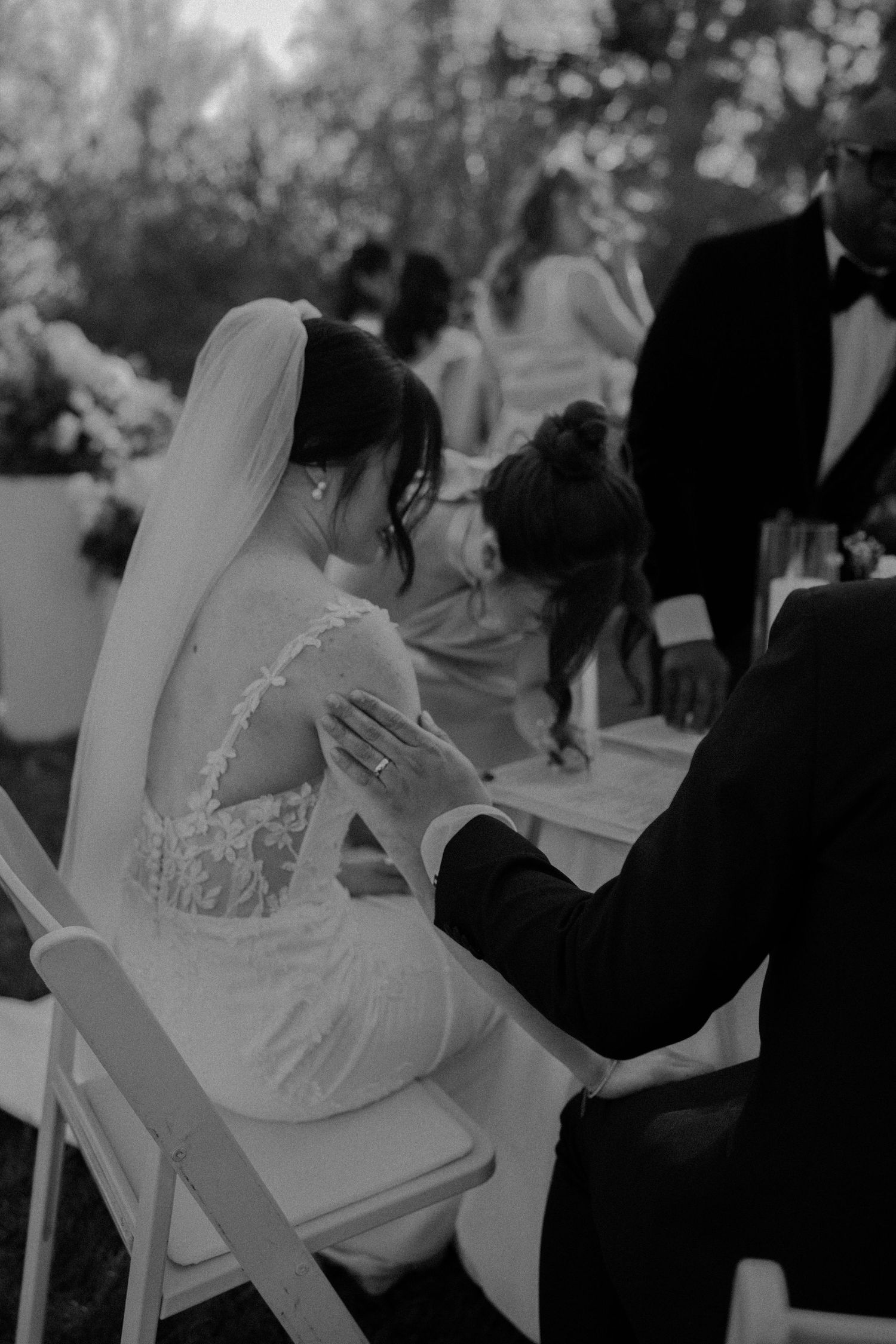 Black and white wedding photo of bride in lace gown with veil, groom placing hand on her back during outdoor ceremony.