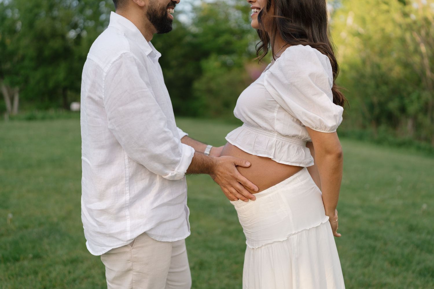 Expecting couple in white outfits holding pregnant belly in a lush green outdoor setting.