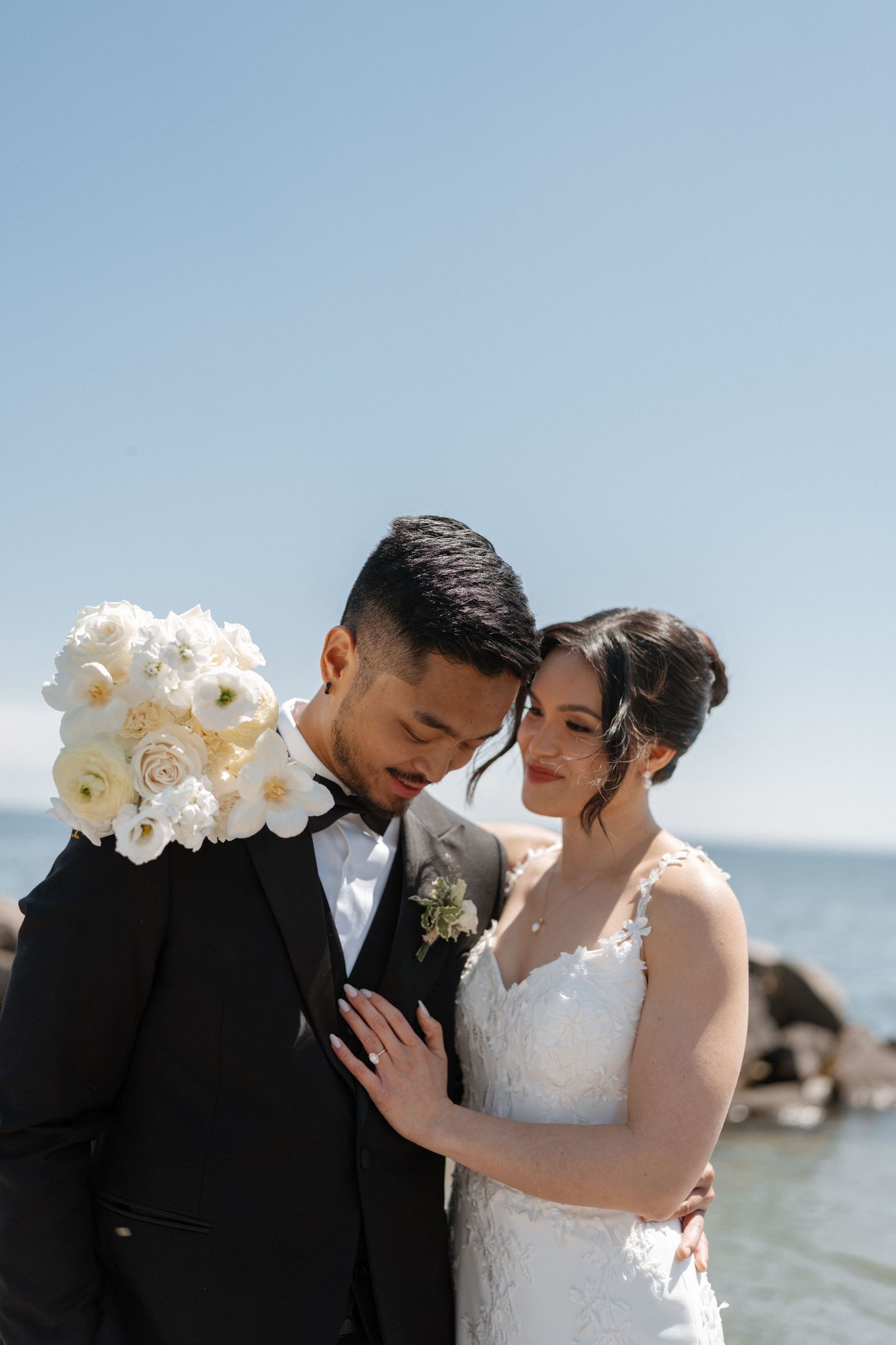 Bride and groom share intimate moment by ocean, she in lace gown holding white roses, he in black tuxedo.