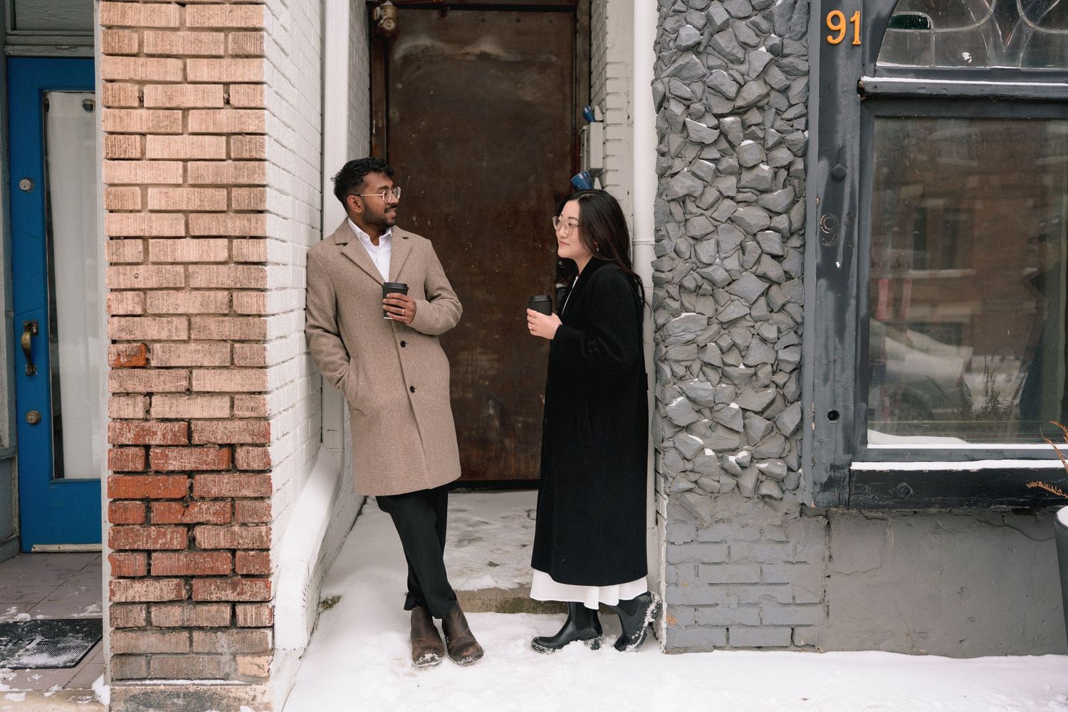 Two people in winter coats having a conversation while standing against a brick wall outside a building.