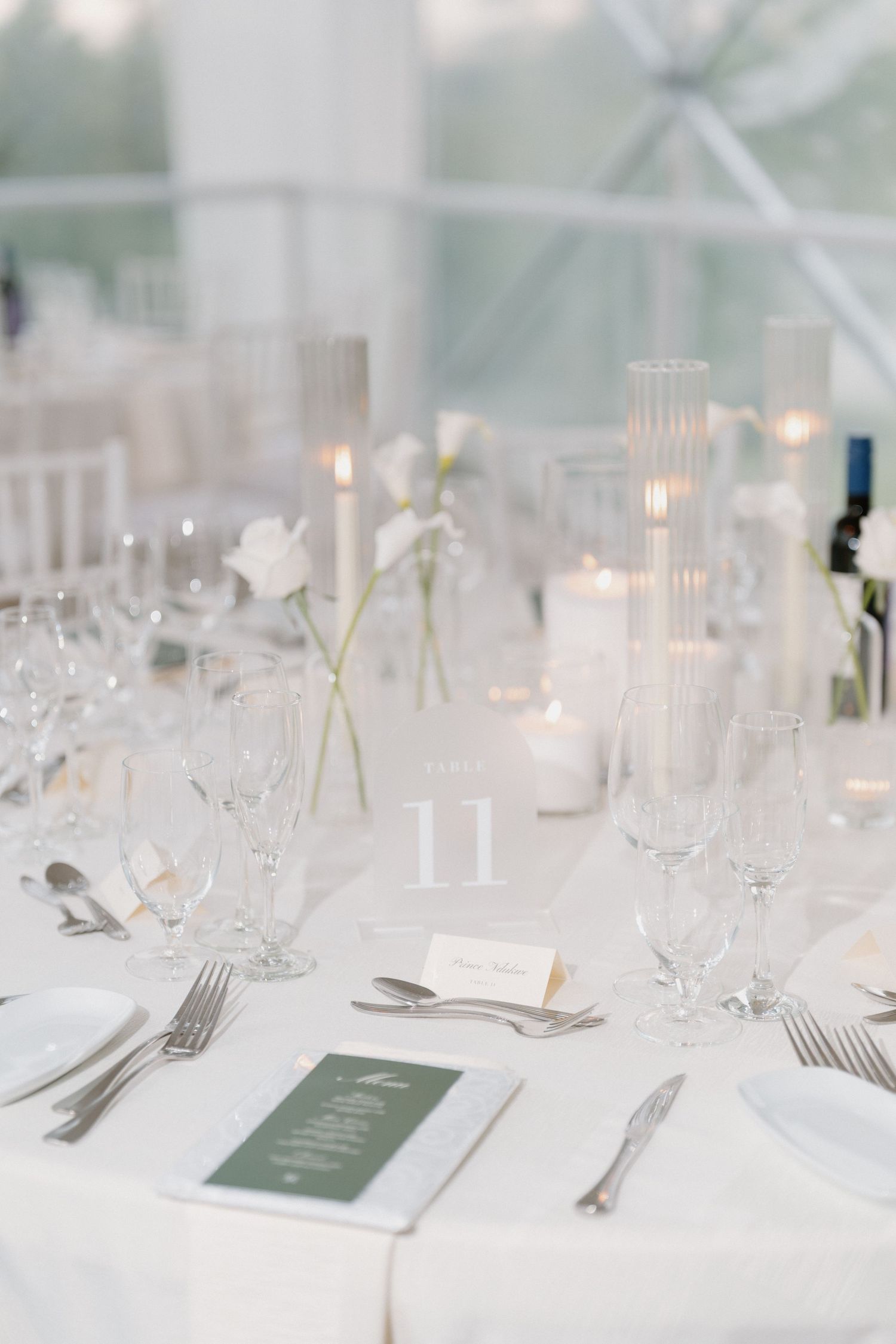 Elegant white wedding reception table setting with crystal glasses, candles, greenery menu cards, and table number 11.