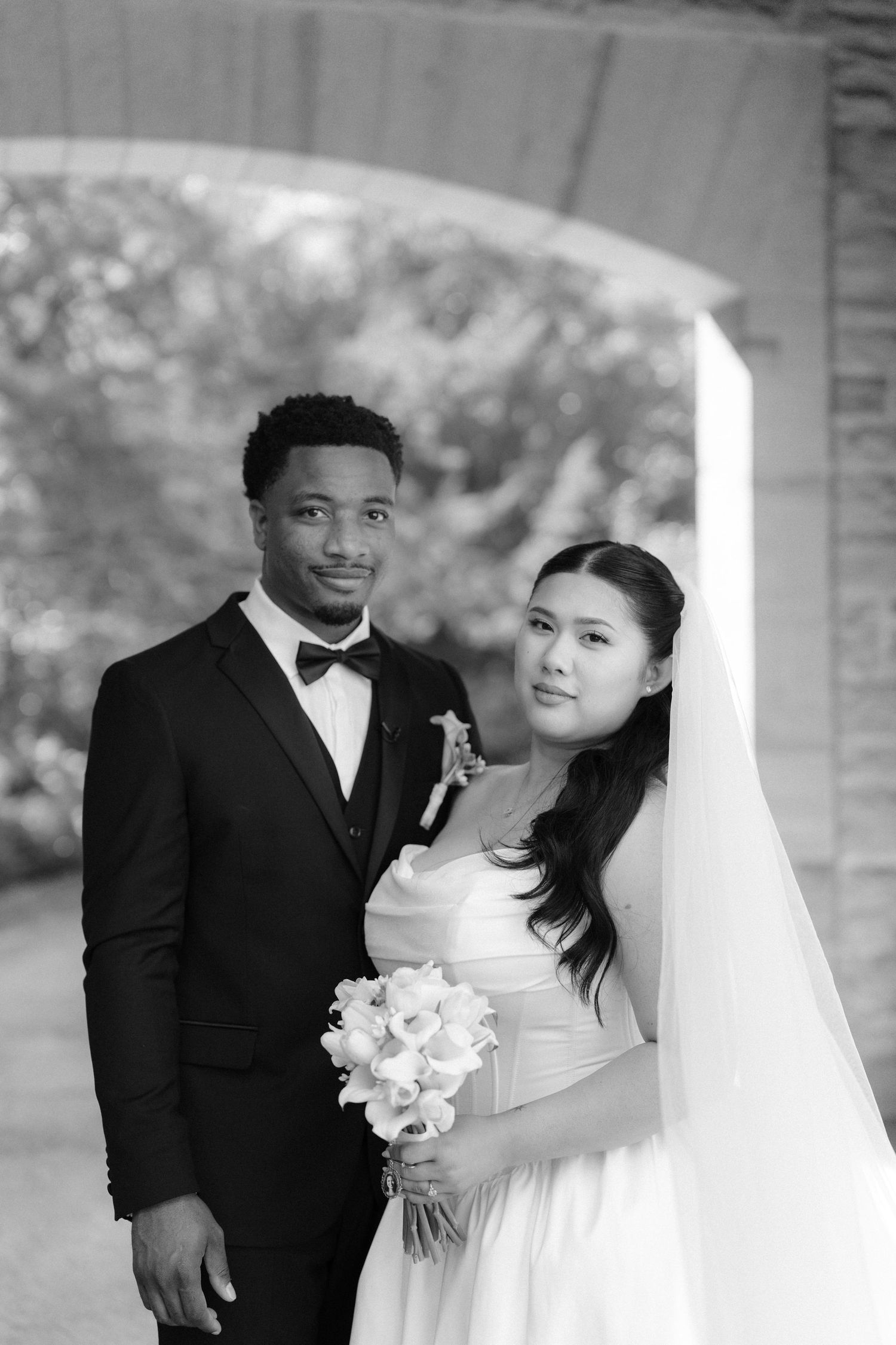 Black and white wedding portrait of a groom in a tuxedo and bride in a white gown holding a bouquet outdoors.