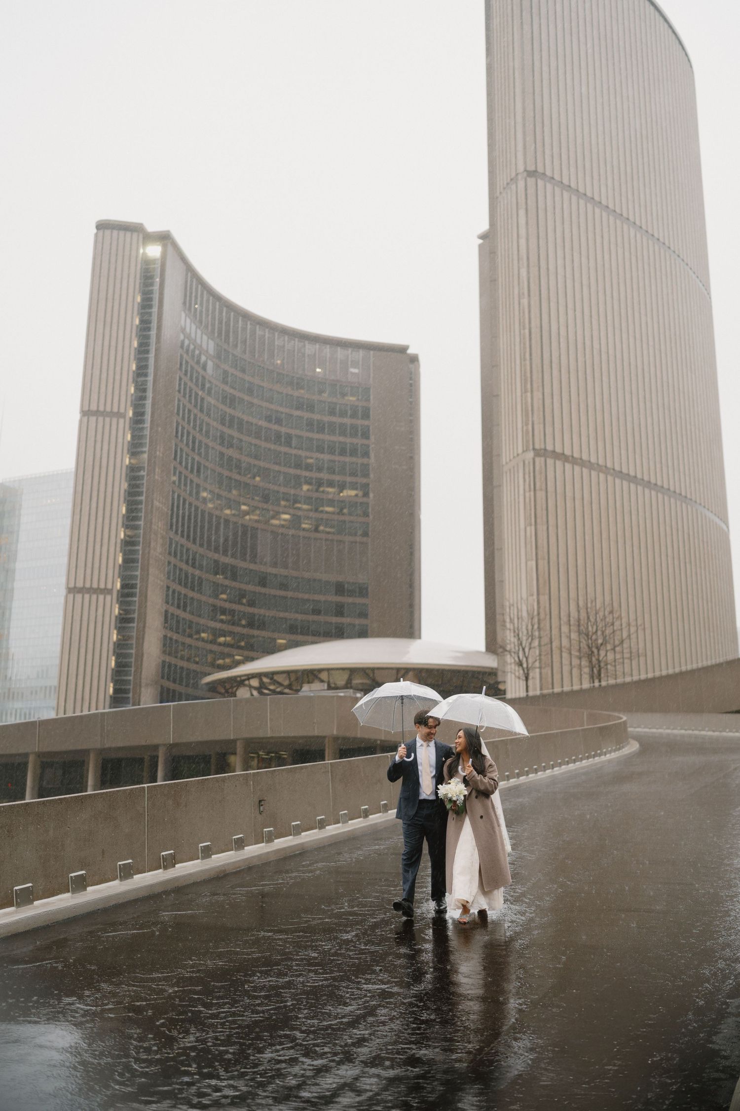Couple sharing umbrella in rain at Toronto City Hall on wedding day.