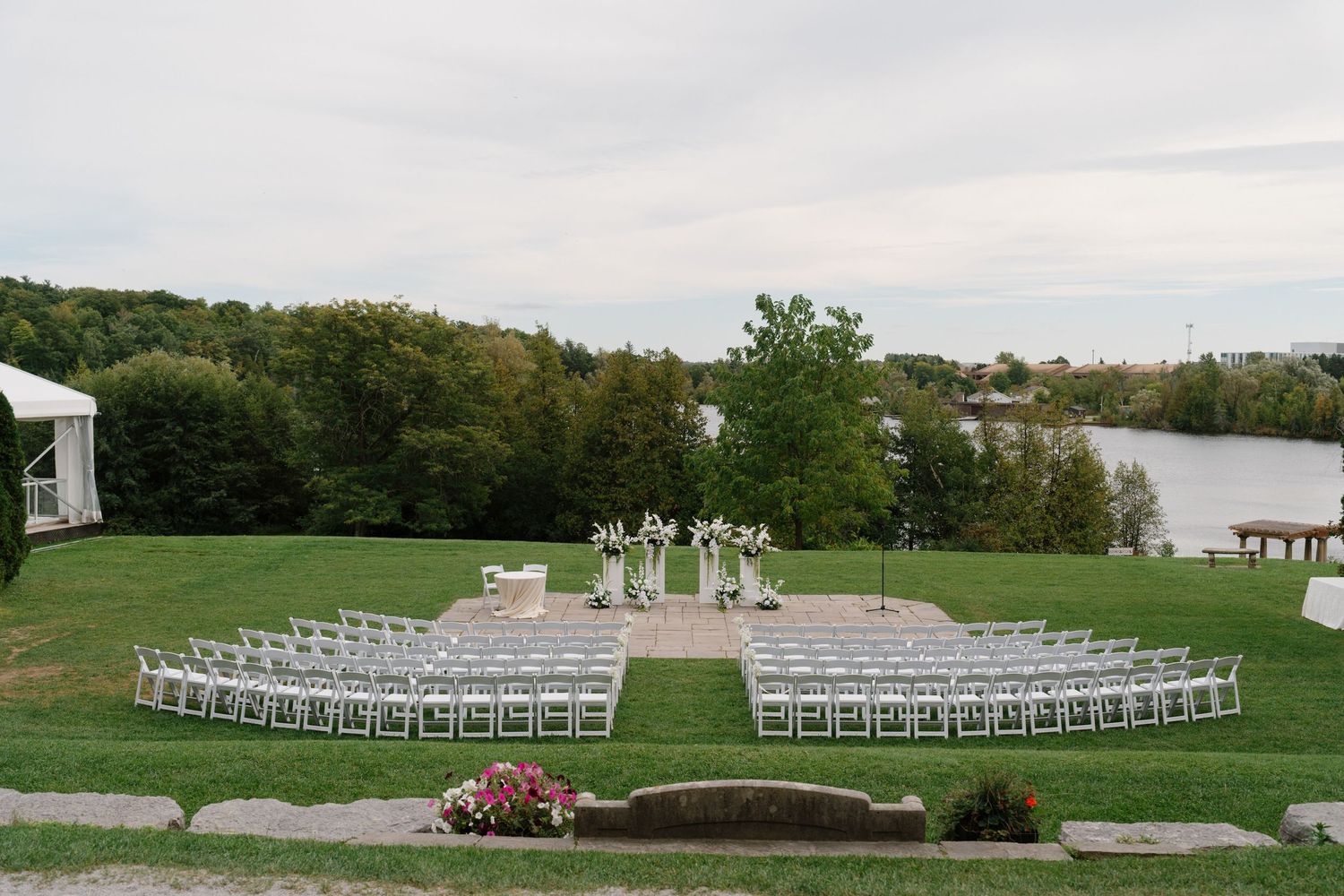 Outdoor wedding ceremony setup with white chairs on green lawn overlooking a serene lake surrounded by lush trees.