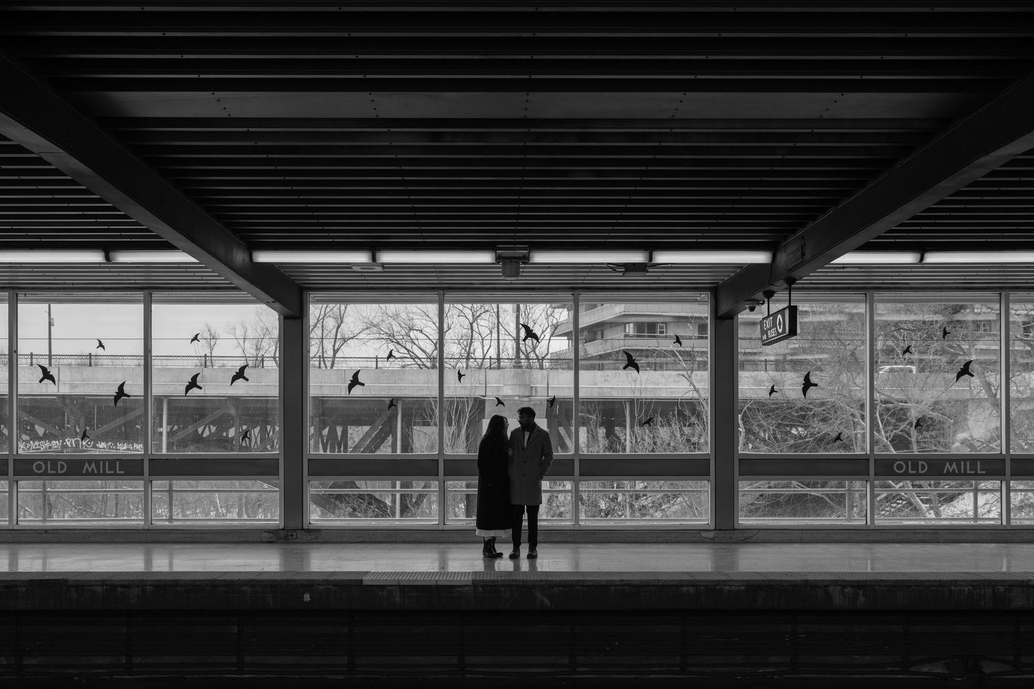 Two silhouettes stand at a train platform with large windows overlooking an urban landscape in black and white.