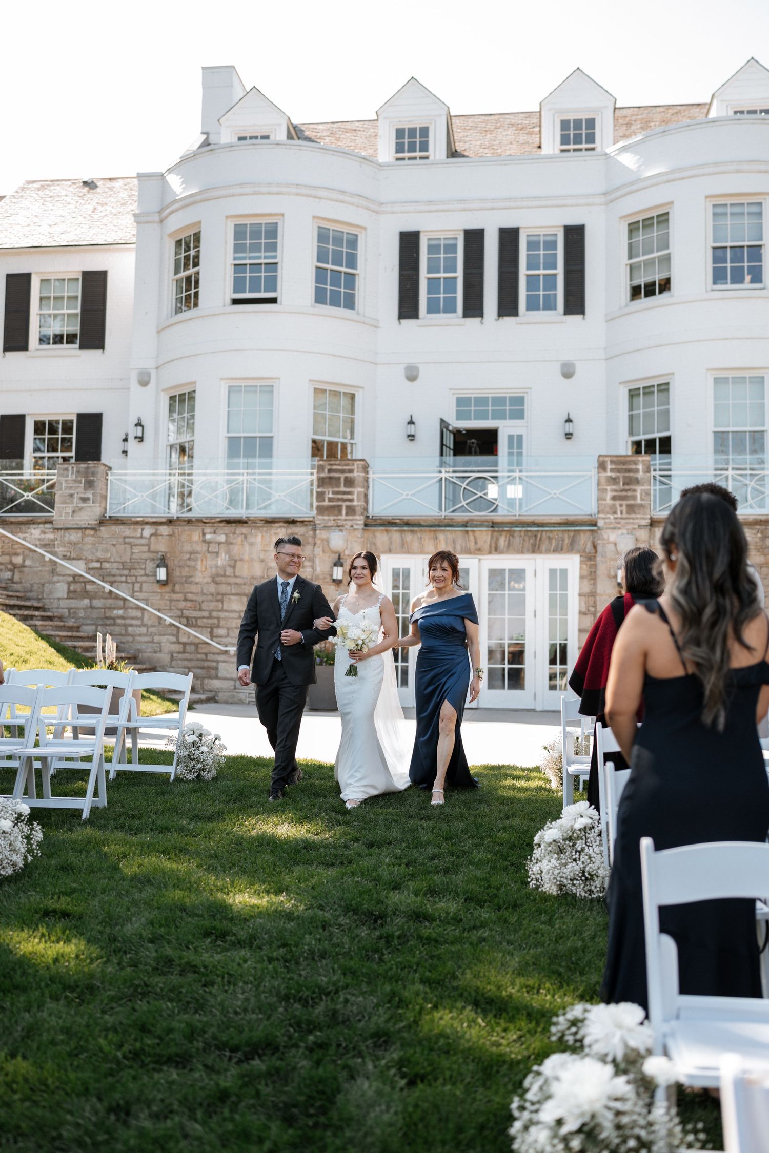 Elegant wedding ceremony scene outside a grand white mansion with bay windows and stone steps on a sunny day.