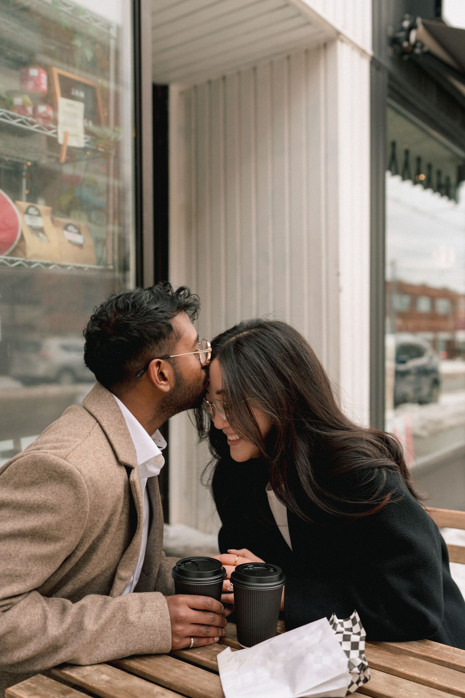 A couple shares an intimate moment while sitting at an outdoor cafe table with coffee cups.
