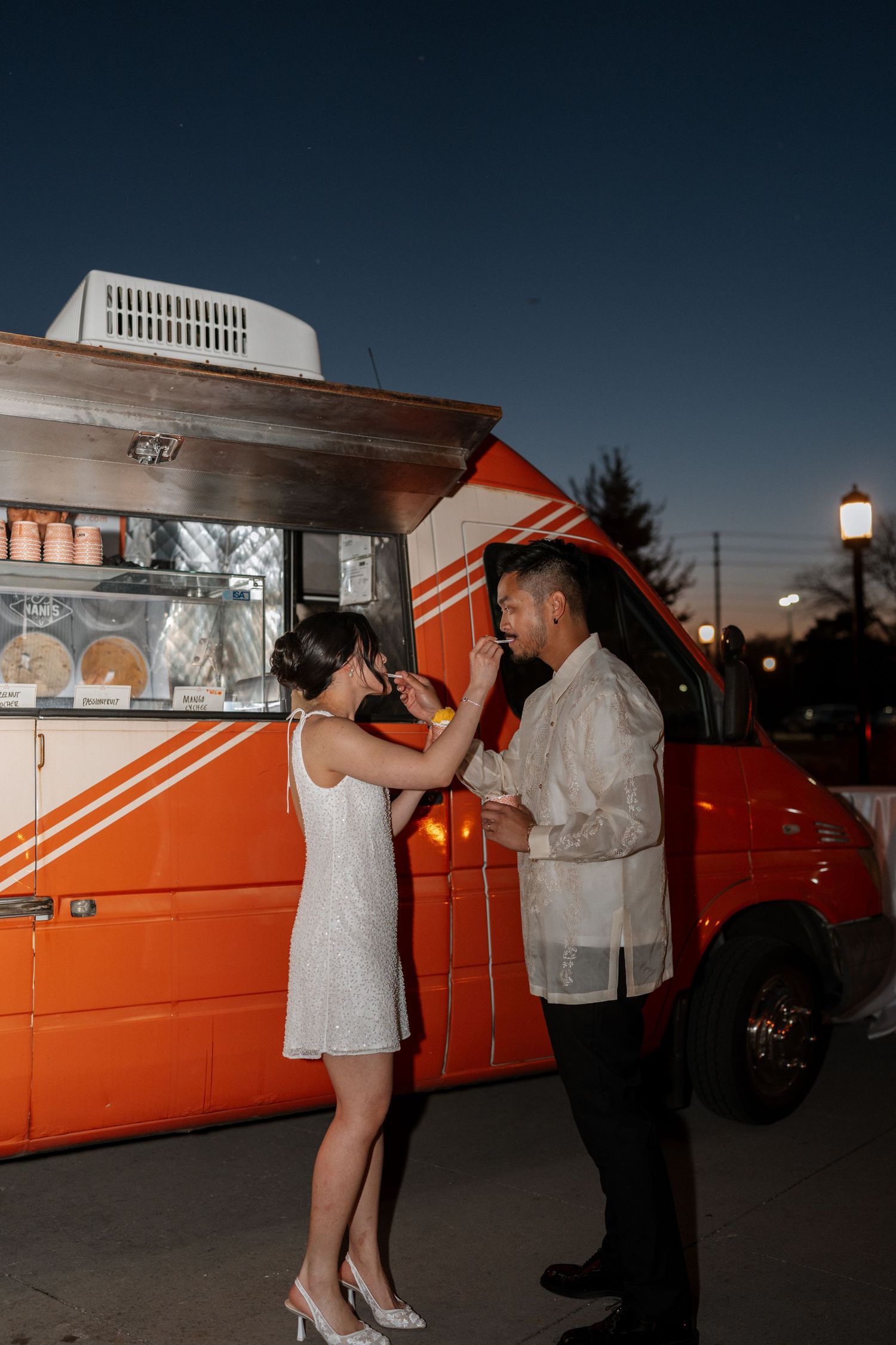A couple shares an intimate moment at dusk in front of a bright orange vintage food truck.
