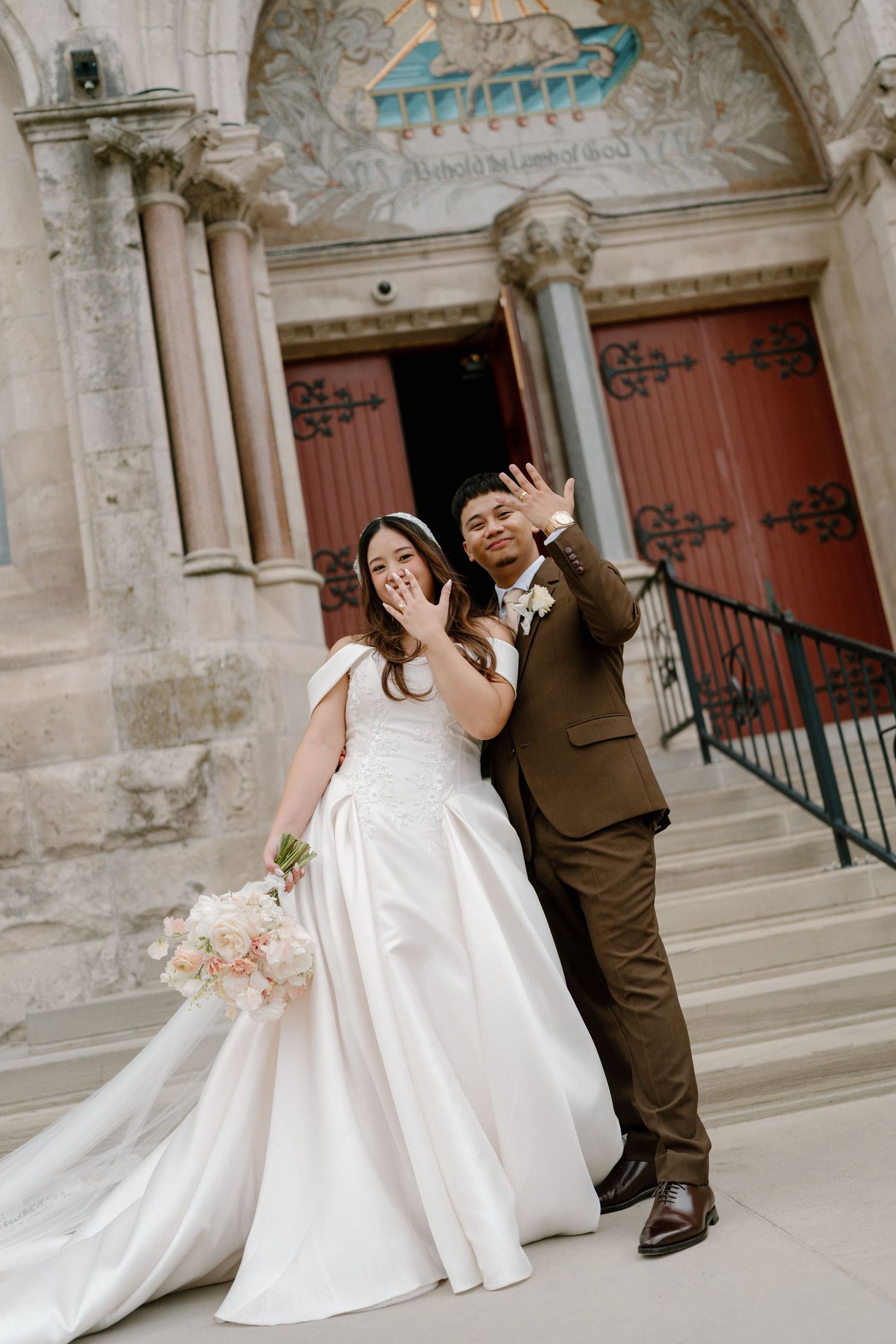 Newlywed couple waving and showing rings outside ornate church, bride in white gown holding bouquet, groom in brown suit.