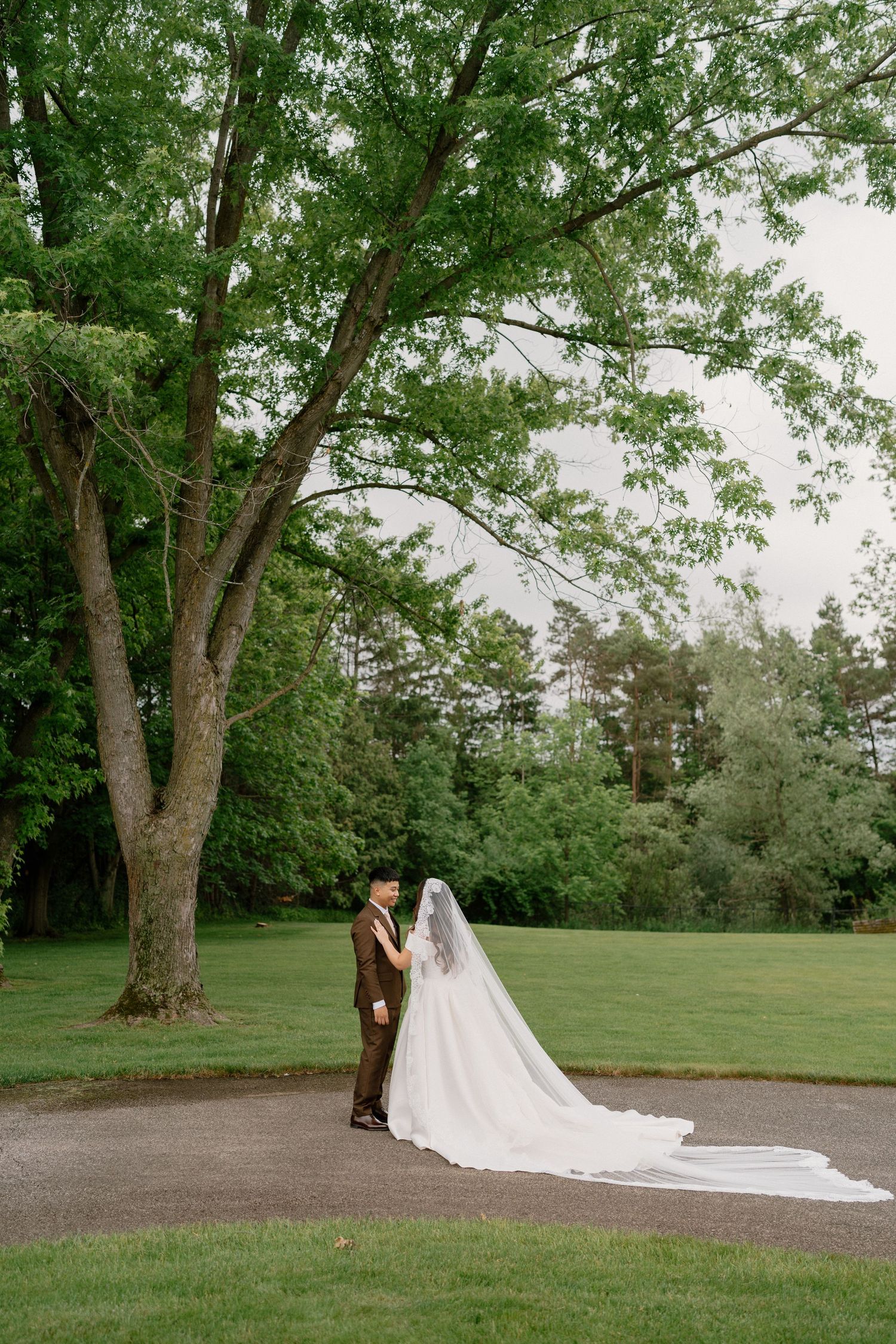 Bride and groom stand beneath a large green tree on a lush lawn, her long white veil trailing on the path.