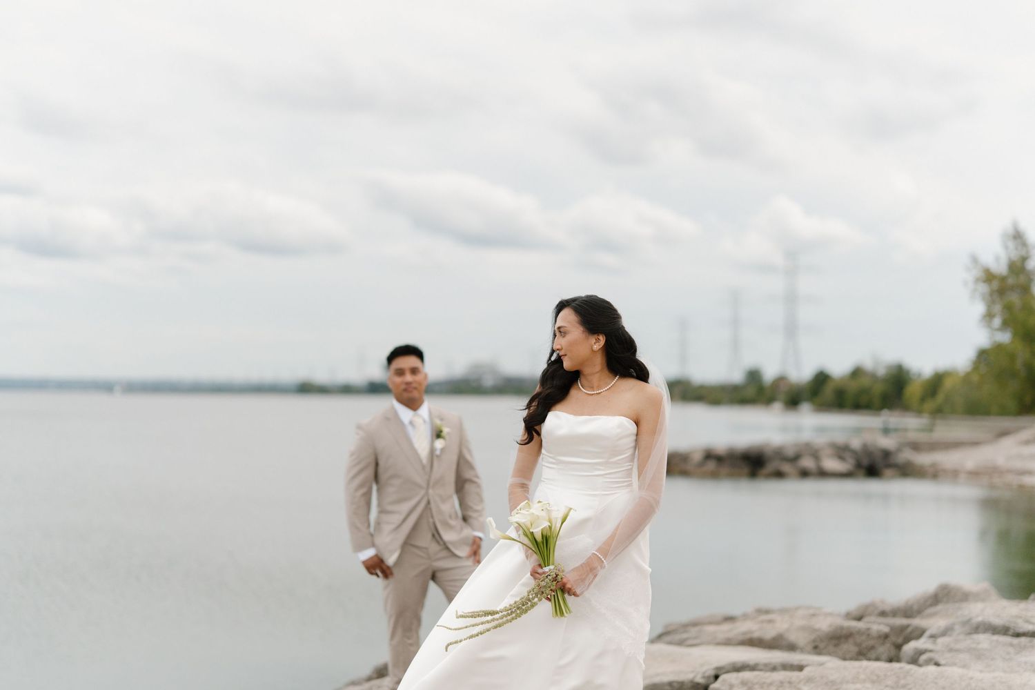 Bride in white strapless gown holds bouquet near waterfront as groom in tan suit stands behind her.