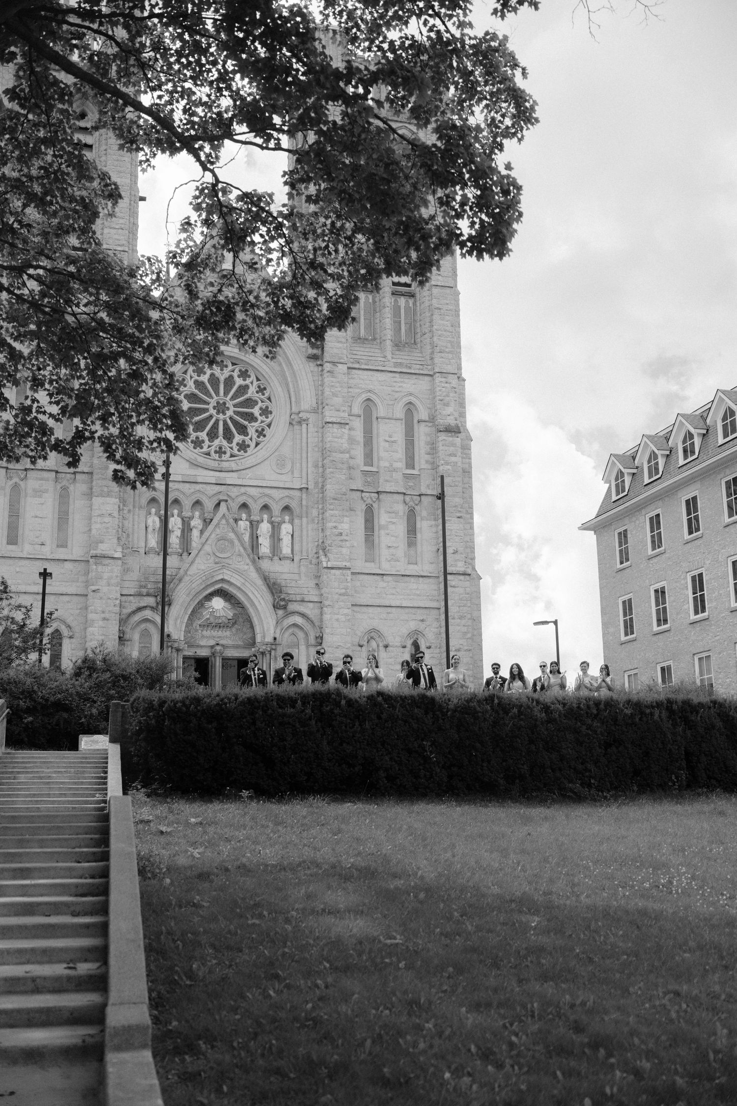 Black and white photo of a Gothic stone cathedral with rose window, surrounded by hedges and trees, with people gathered outside.