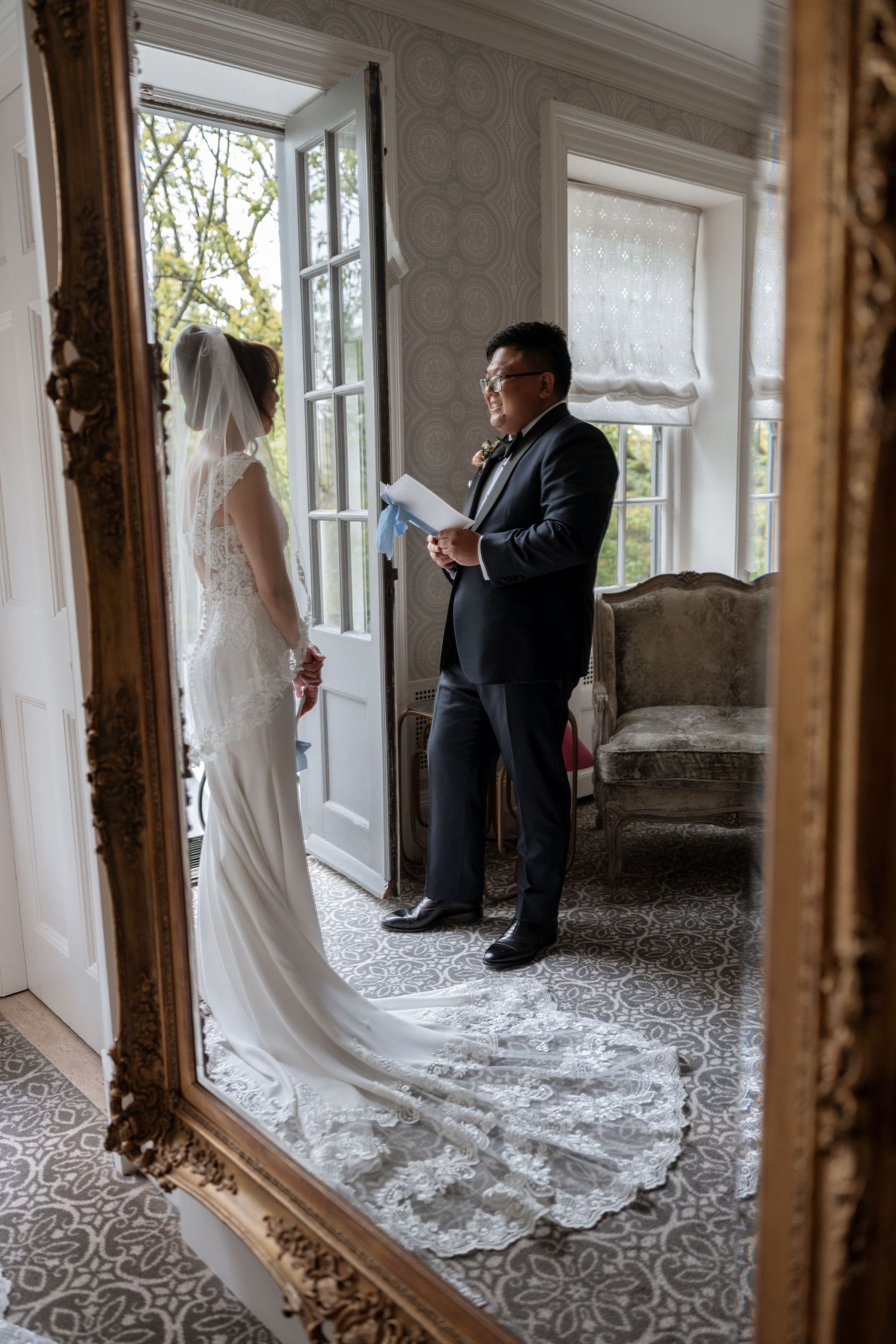 Wedding ceremony reflection in ornate gold mirror shows officiant and bride in elegant white gown by bright window.