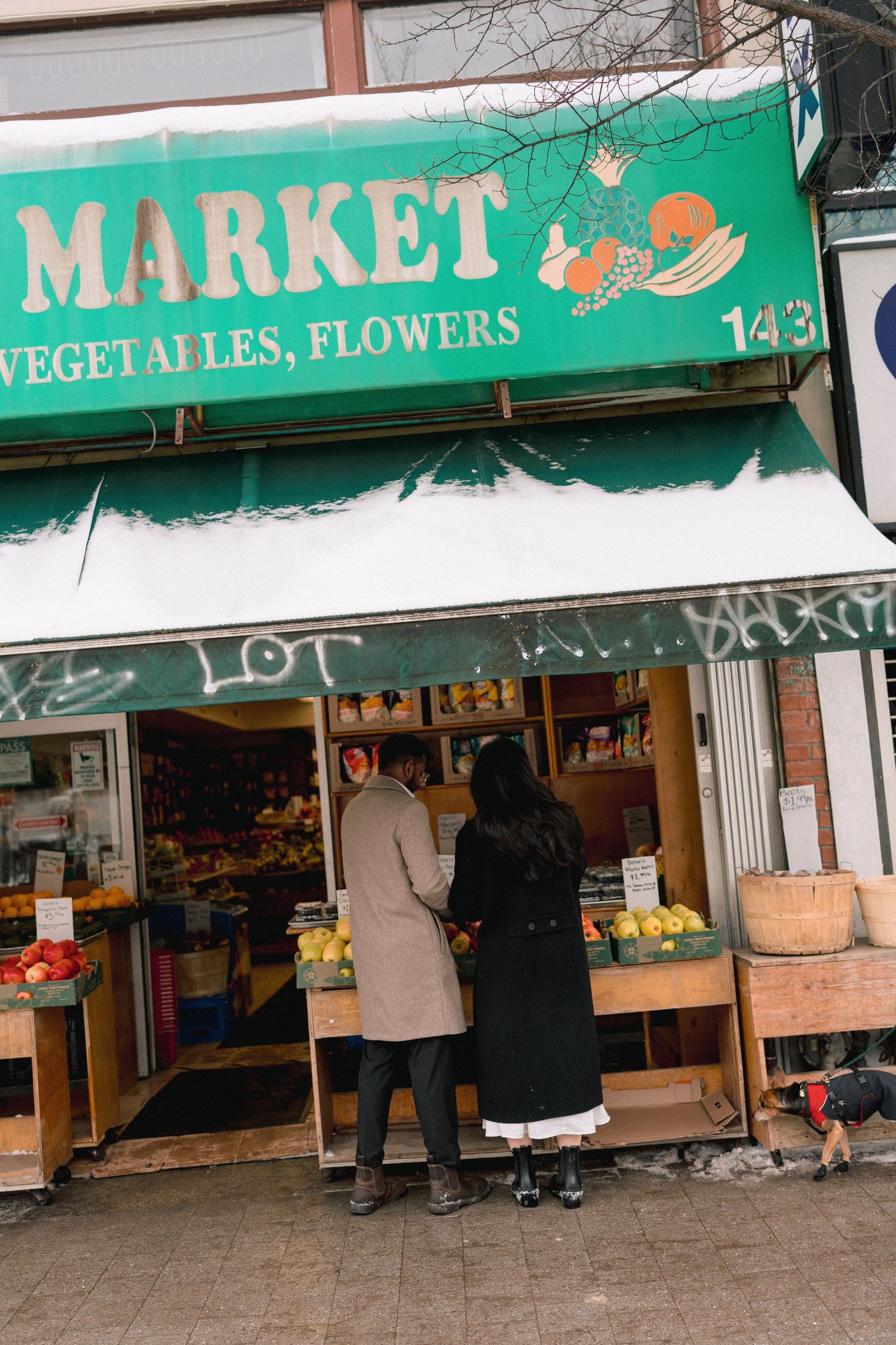 Exterior view of a local market with green awning displaying 'Market' and 'Vegetables, Flowers' signage with customers shopping.