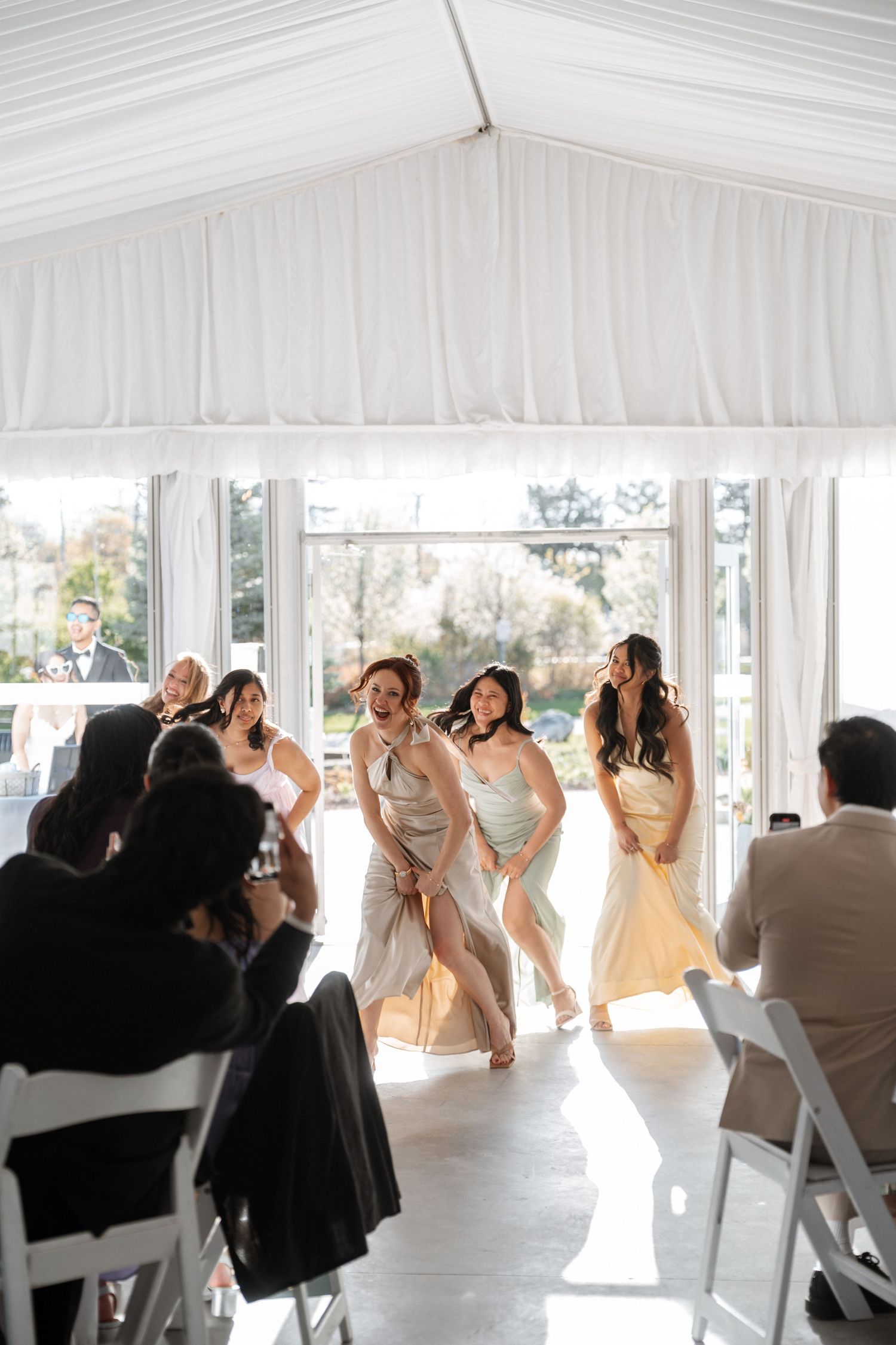 Bridesmaids in flowing champagne dresses dance together in sunlit reception venue with white draping and folding chairs.