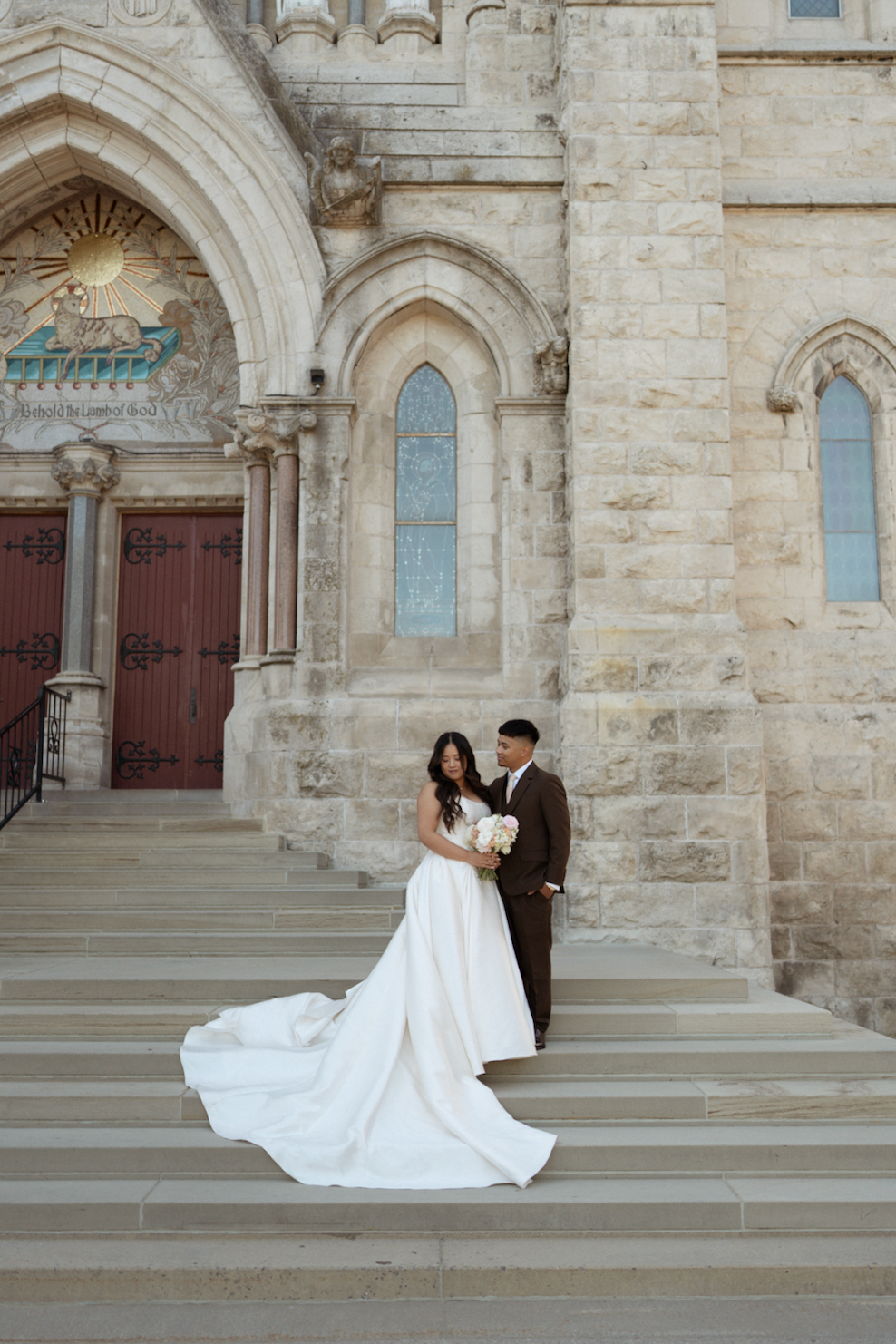 Bride in white gown with long train and groom in brown suit pose on church steps for wedding photos.