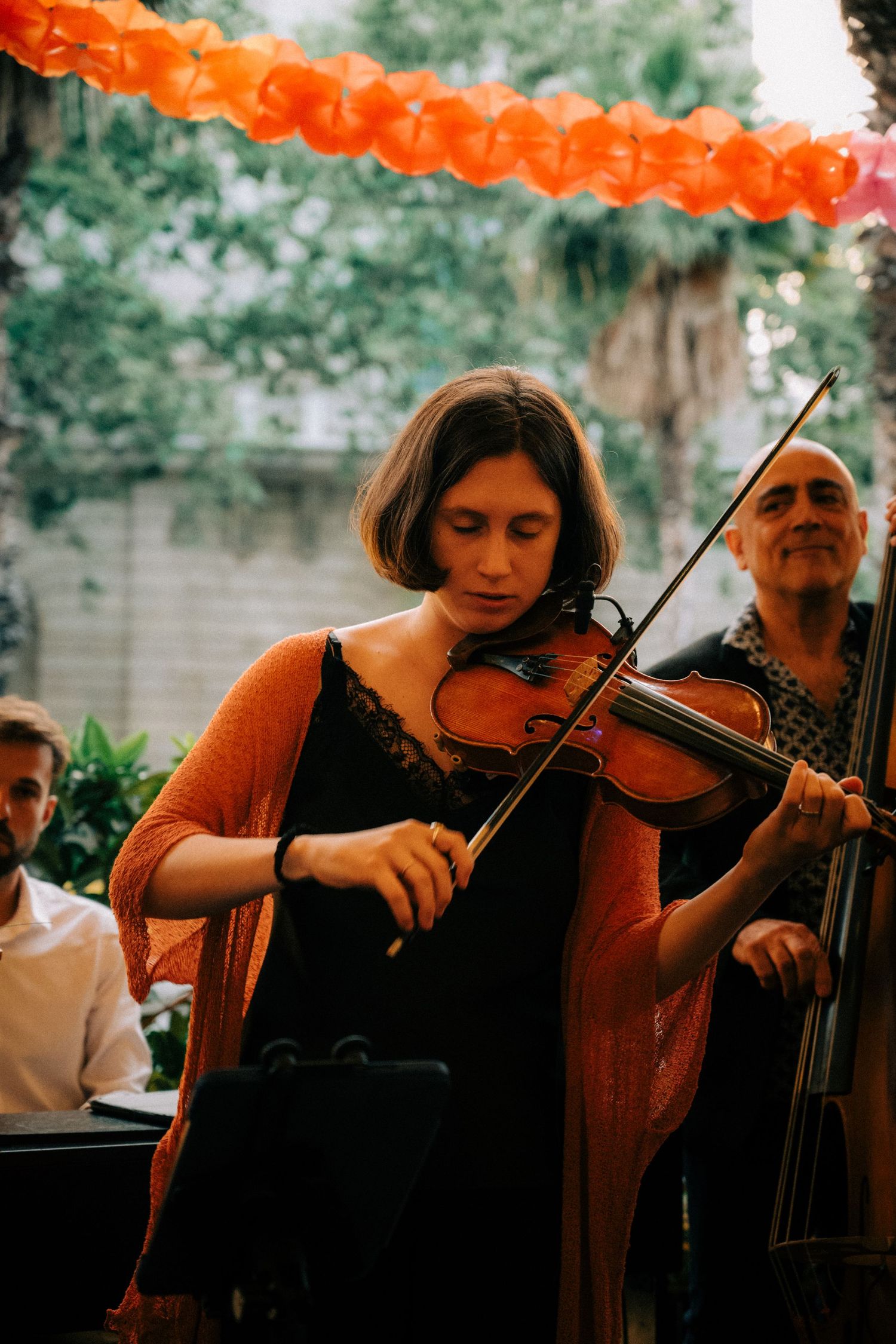 Musicians perform with string instruments under orange paper lantern decorations outdoors.