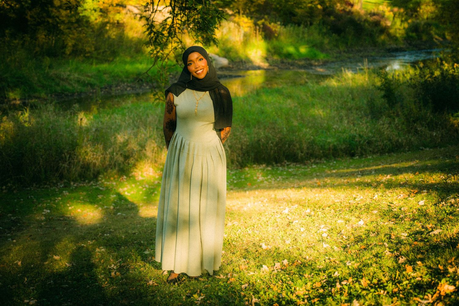 A woman in flowing dress stands in a sun-dappled meadow with golden light.