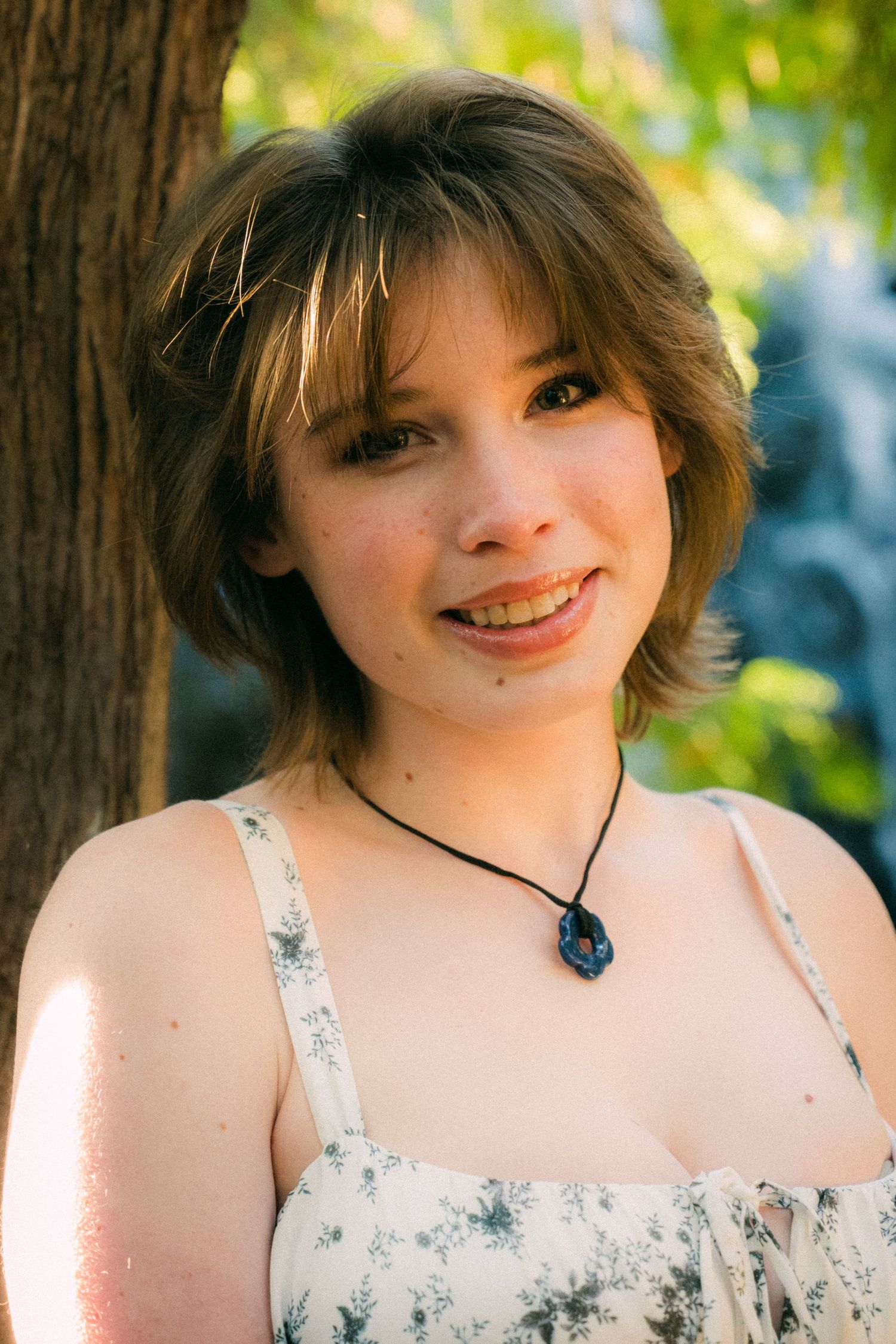 A person in a light summer dress smiles warmly in natural outdoor lighting.