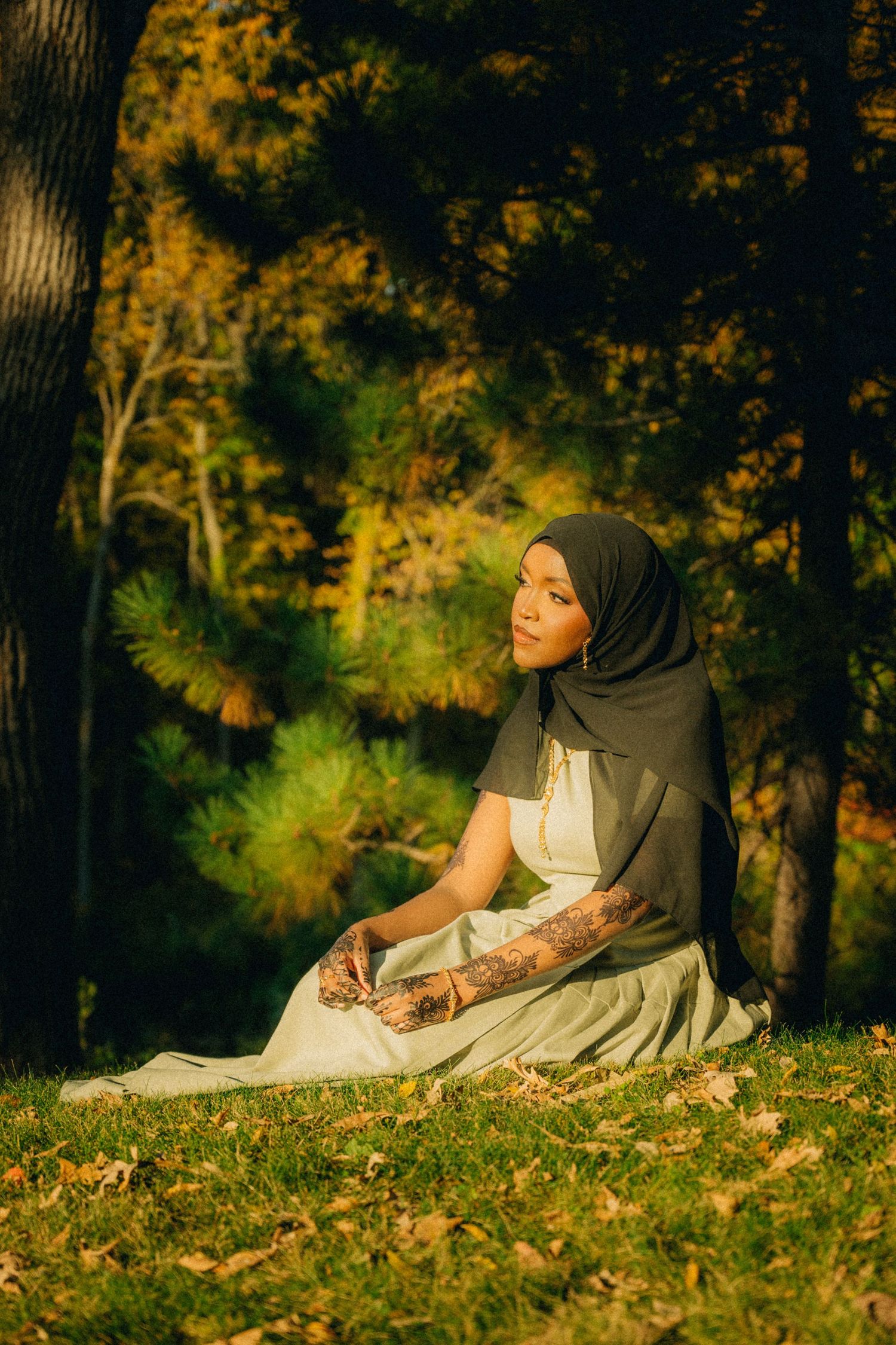 A woman in white dress and dark hijab sits peacefully on an autumn hillside.