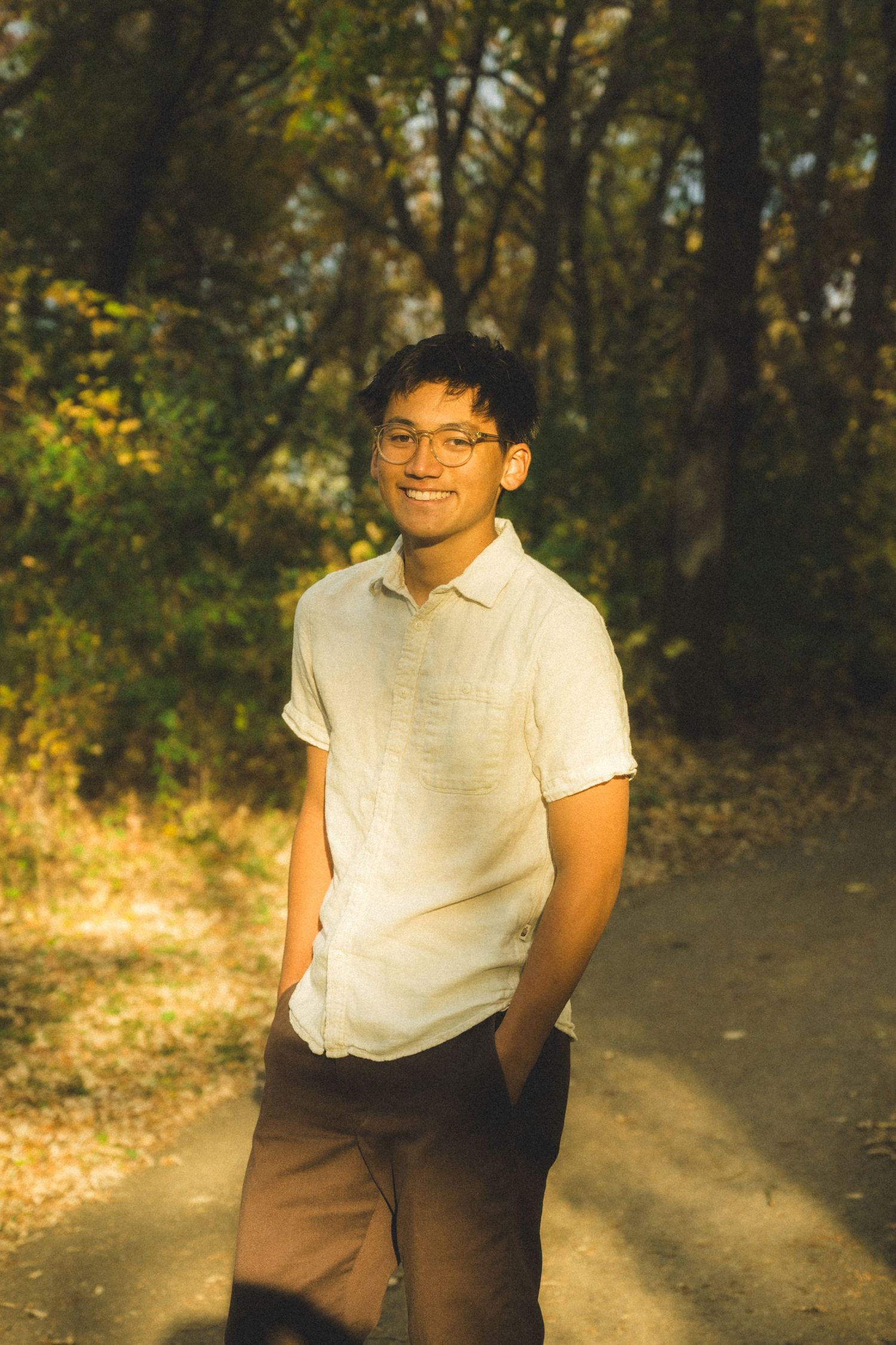 Person in white polo shirt and brown pants standing on wooded path during golden hour.