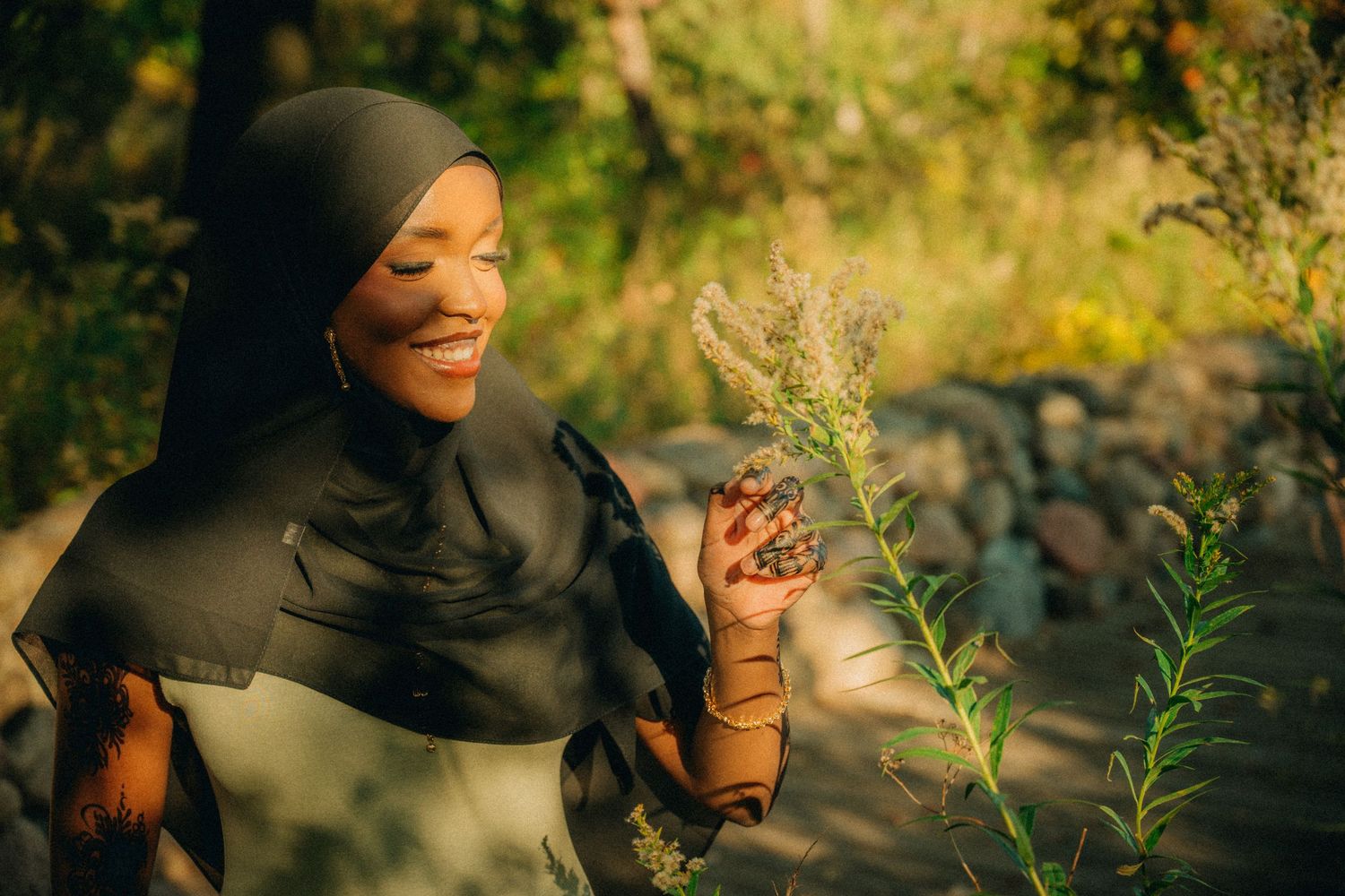 Person in black hijab smiling while touching plants in a sunlit garden setting.