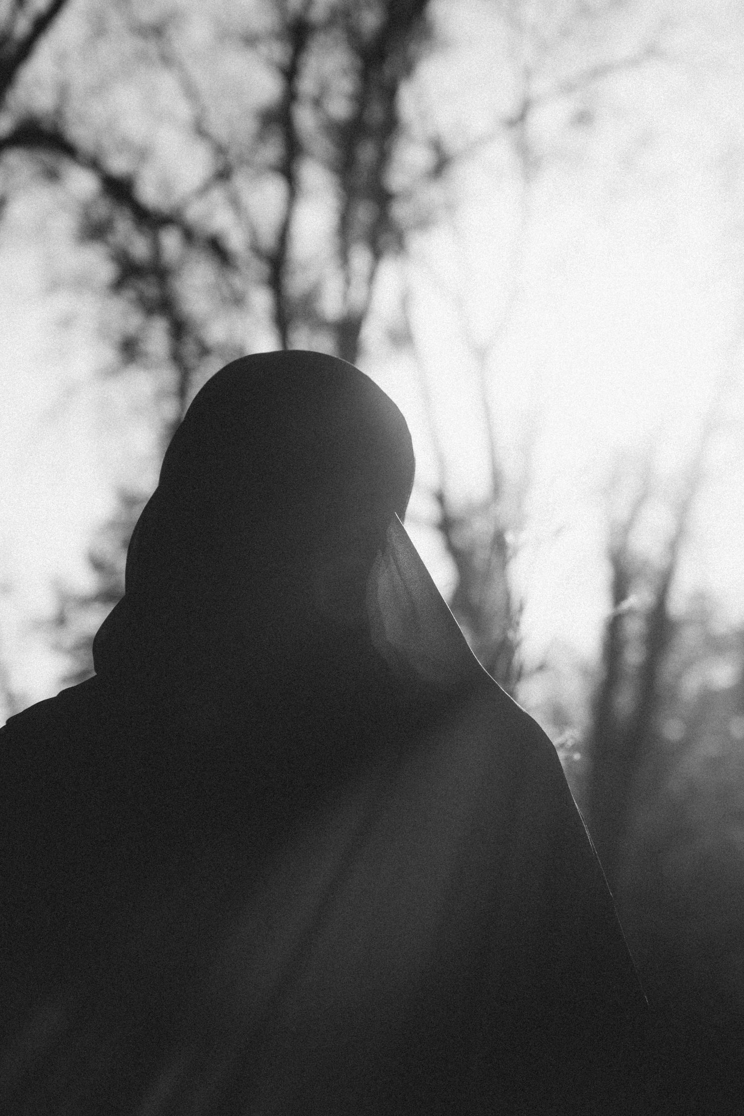 A silhouetted figure in hijab against a bright, hazy sky with bare trees.