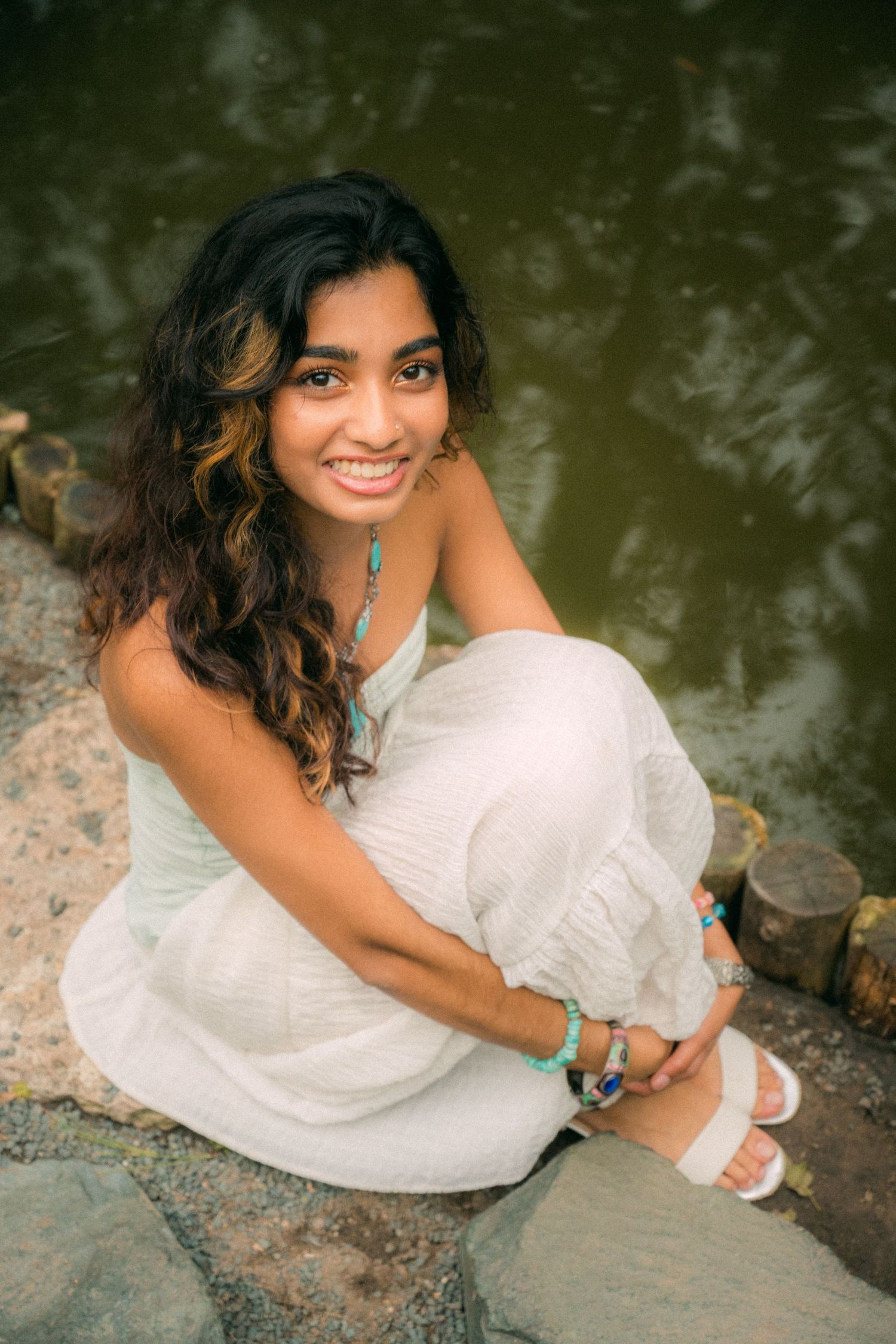 Person in white dress sitting by water's edge during outdoor photo session.