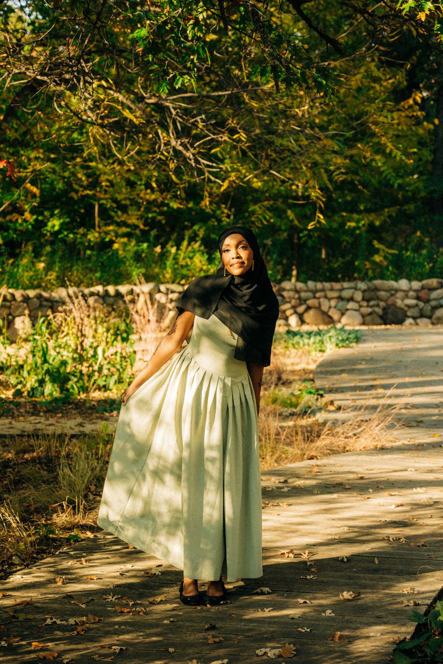 A woman in white dress walks along a sunlit garden path beneath overhanging trees.