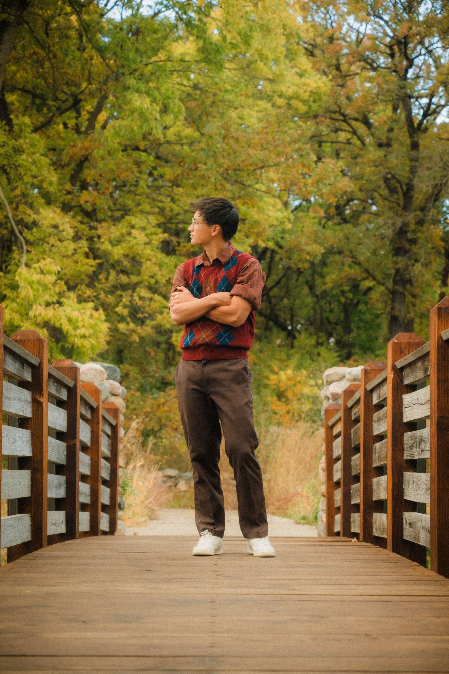 Person in plaid shirt and brown pants standing confidently on wooden bridge during autumn.
