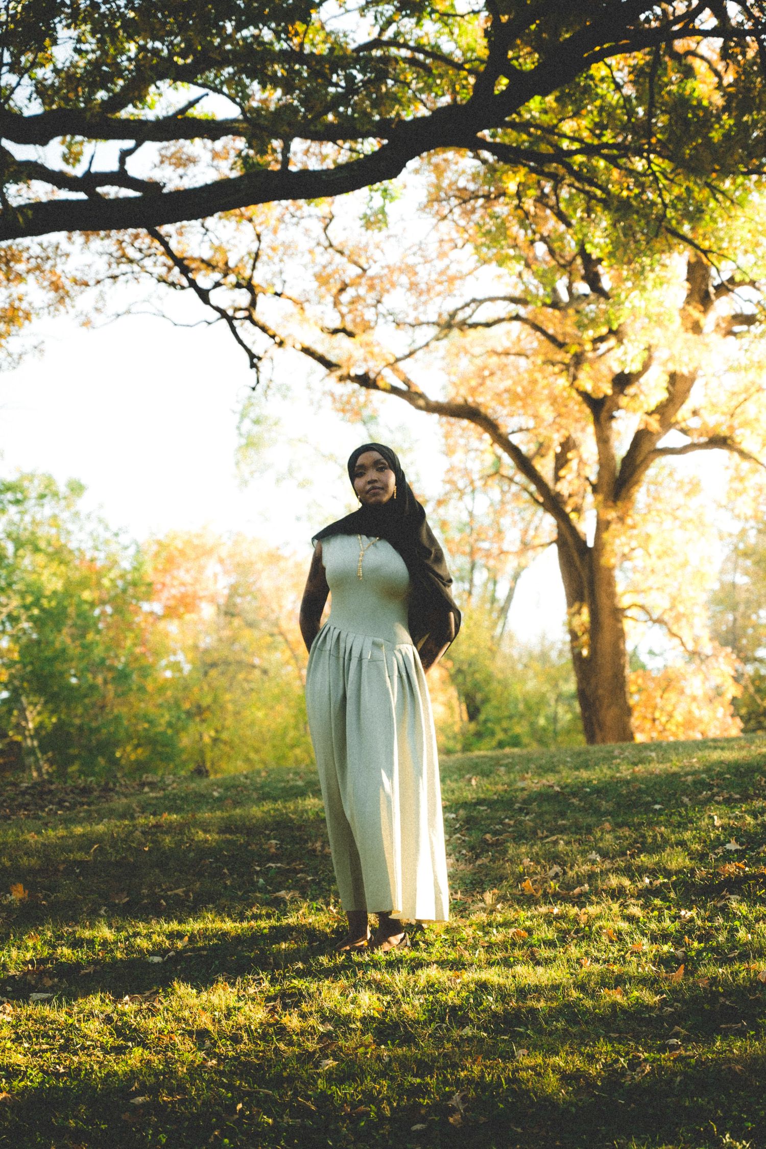 A woman in traditional dress stands beneath autumn trees in golden afternoon light.