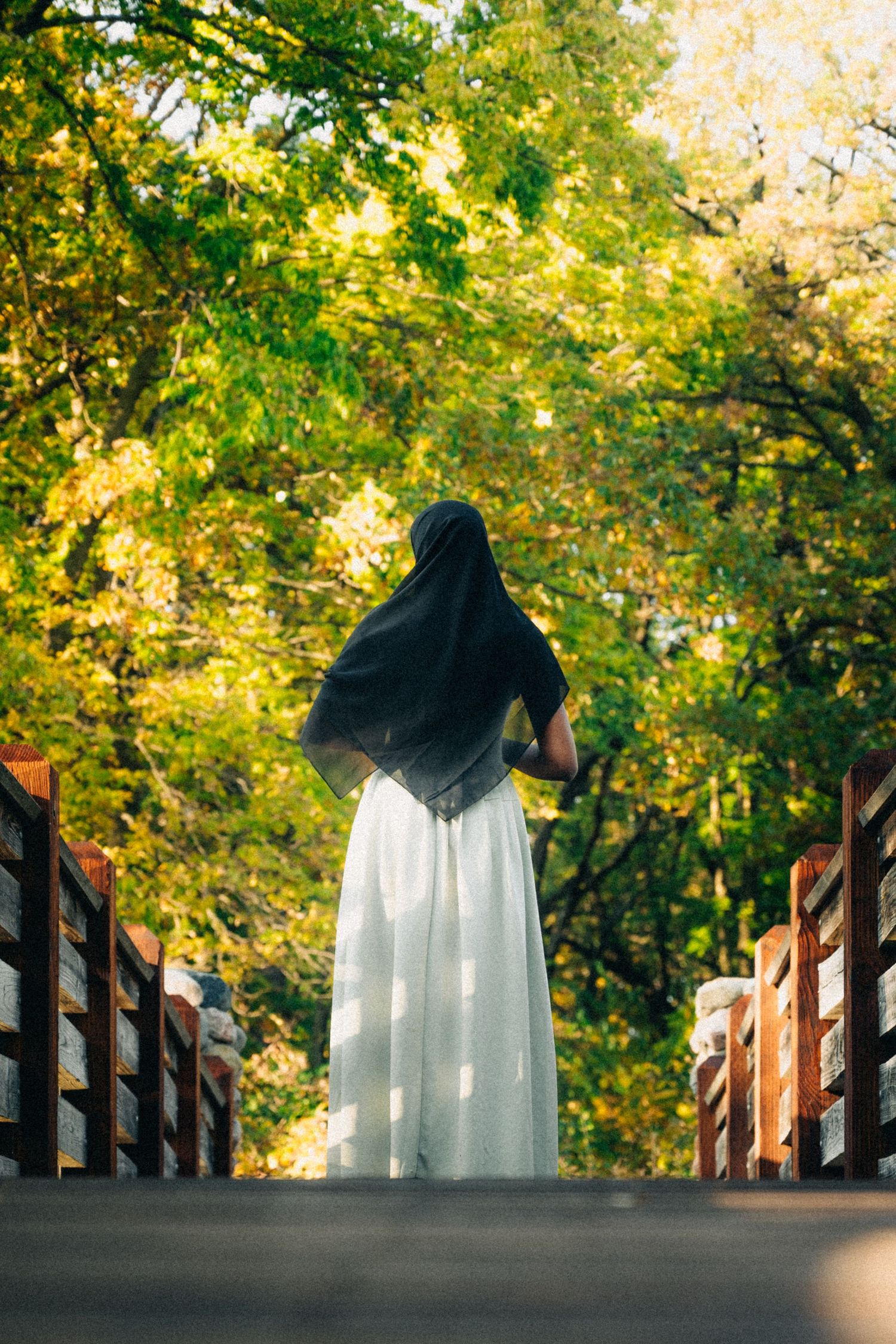 A figure in white dress and black hijab stands on a wooden bridge surrounded by autumn foliage.