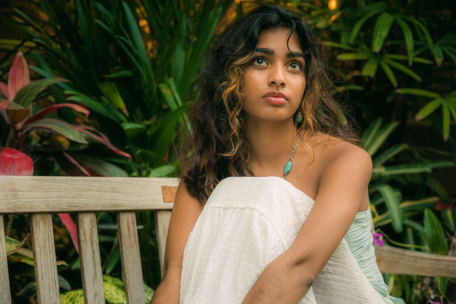 A portrait of someone in a white dress sitting on a wooden bench with tropical plants in the background.