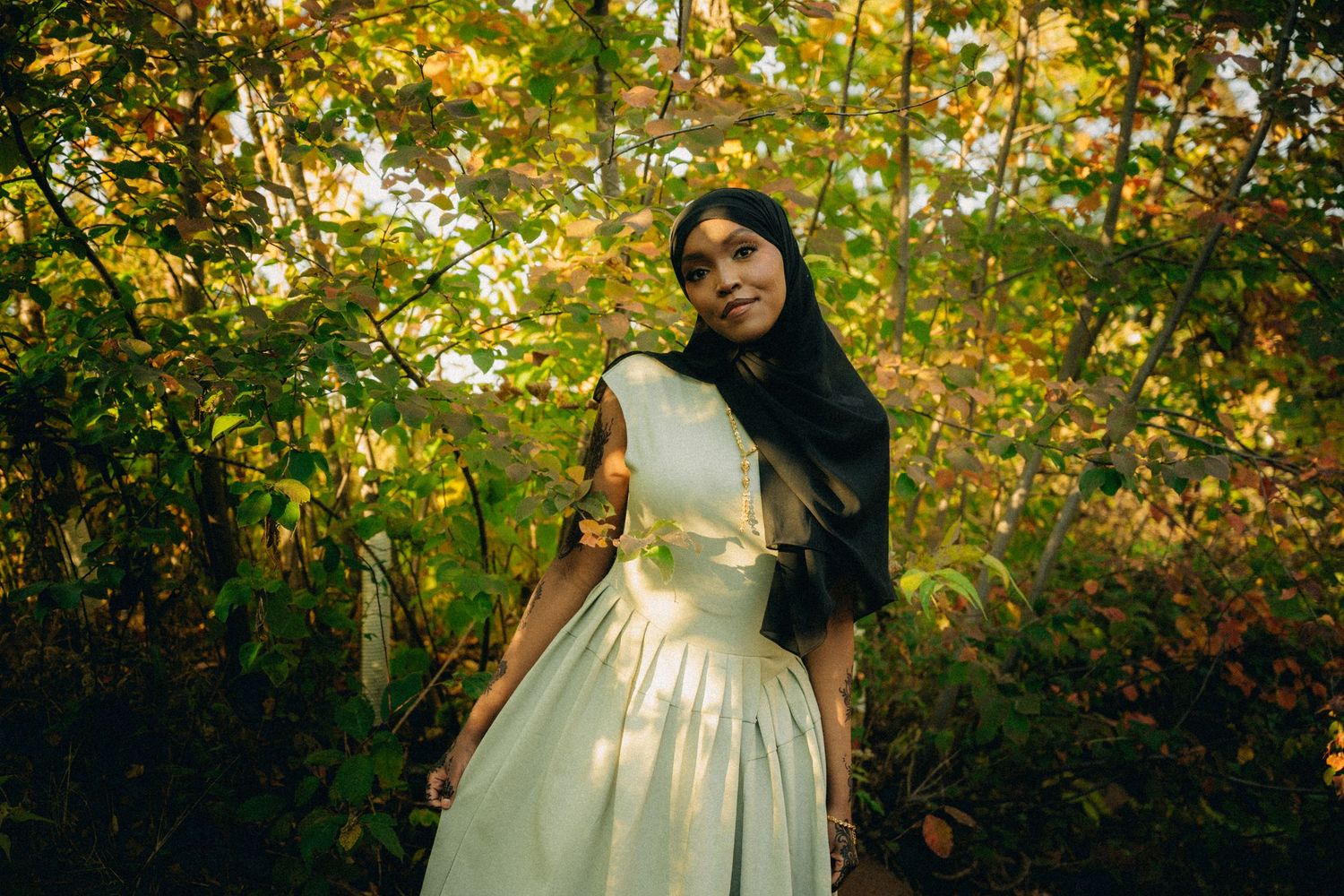 A woman in a white dress and black hijab poses in a sunlit autumn forest setting.