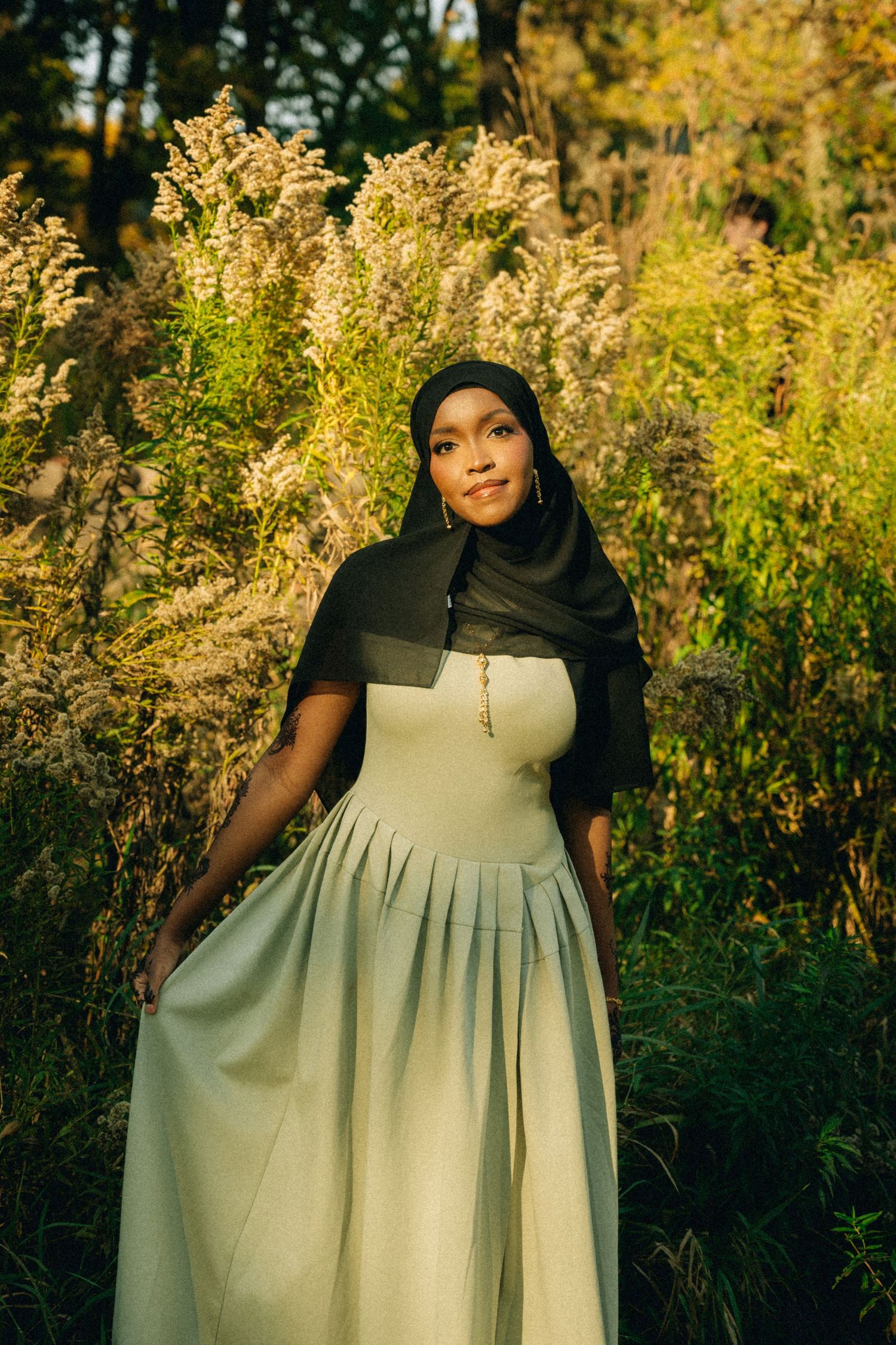 Woman in light-colored dress and black hijab poses in front of tall golden grasses at sunset.