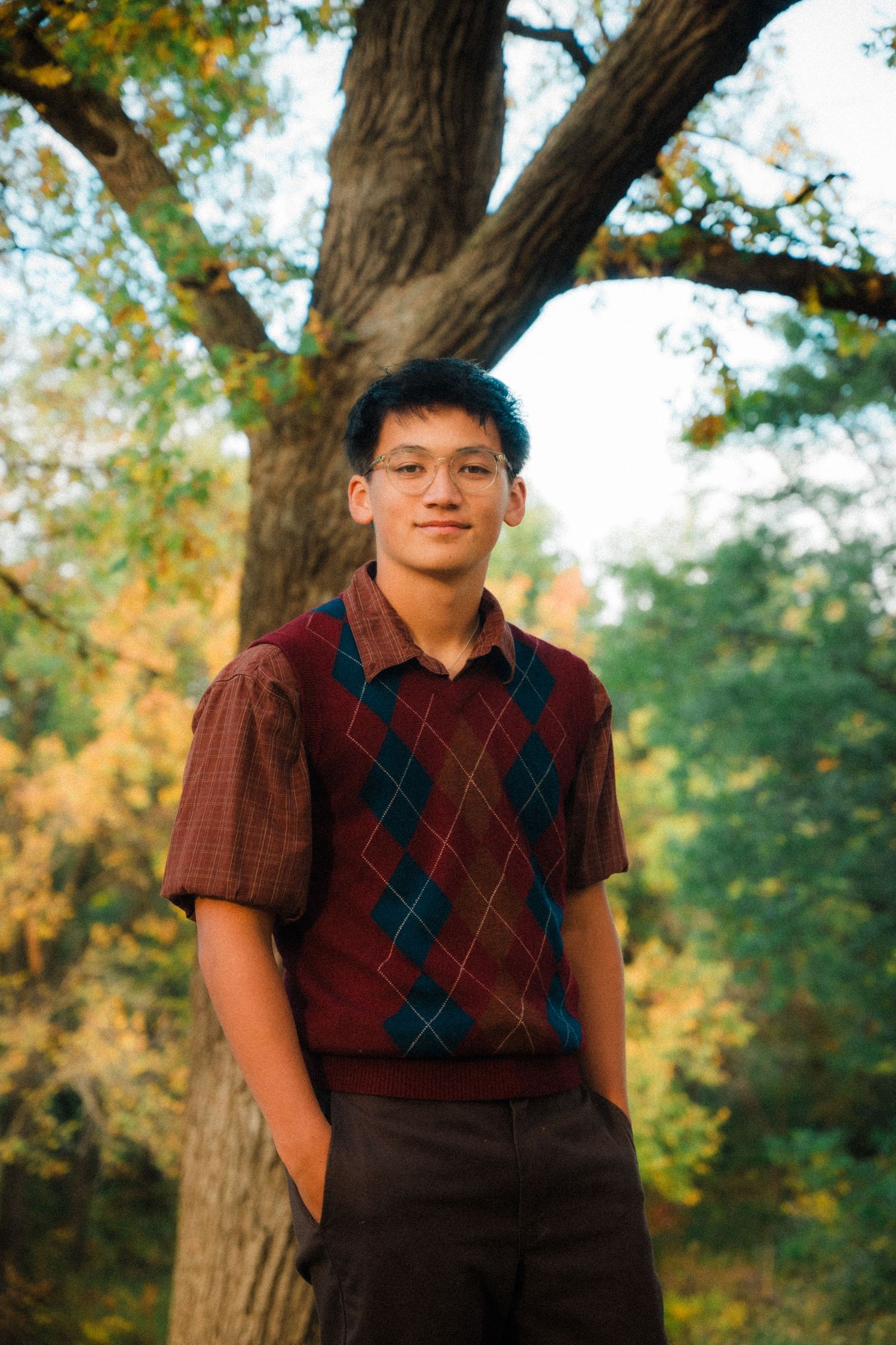 Person in burgundy argyle sweater vest posing in front of fall foliage.