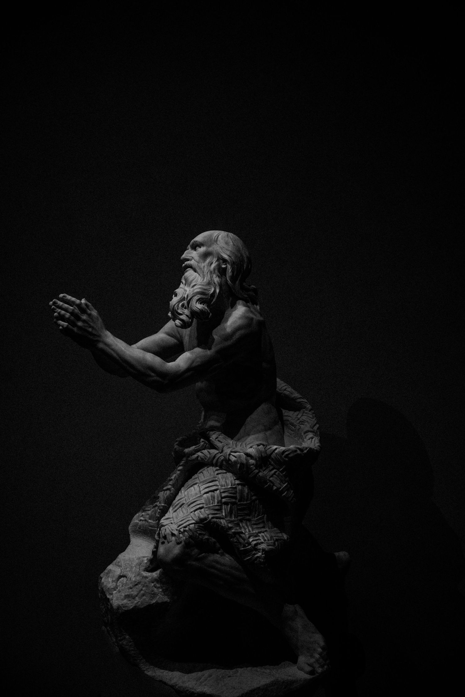 Dramatic black and white sculpture of a bearded figure reaching upward against dark background.