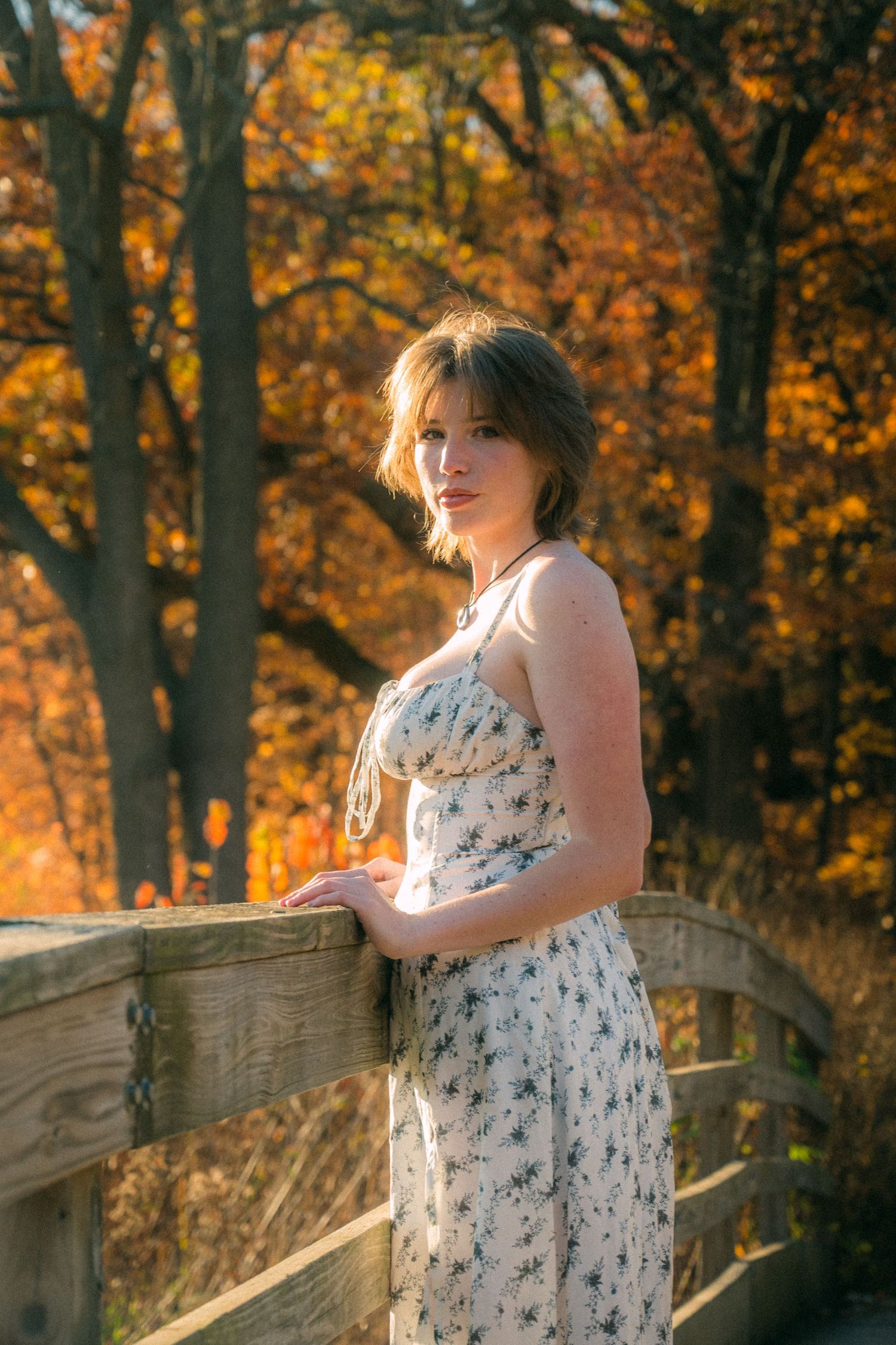 A person in a white floral dress poses on a wooden bridge surrounded by autumn foliage.
