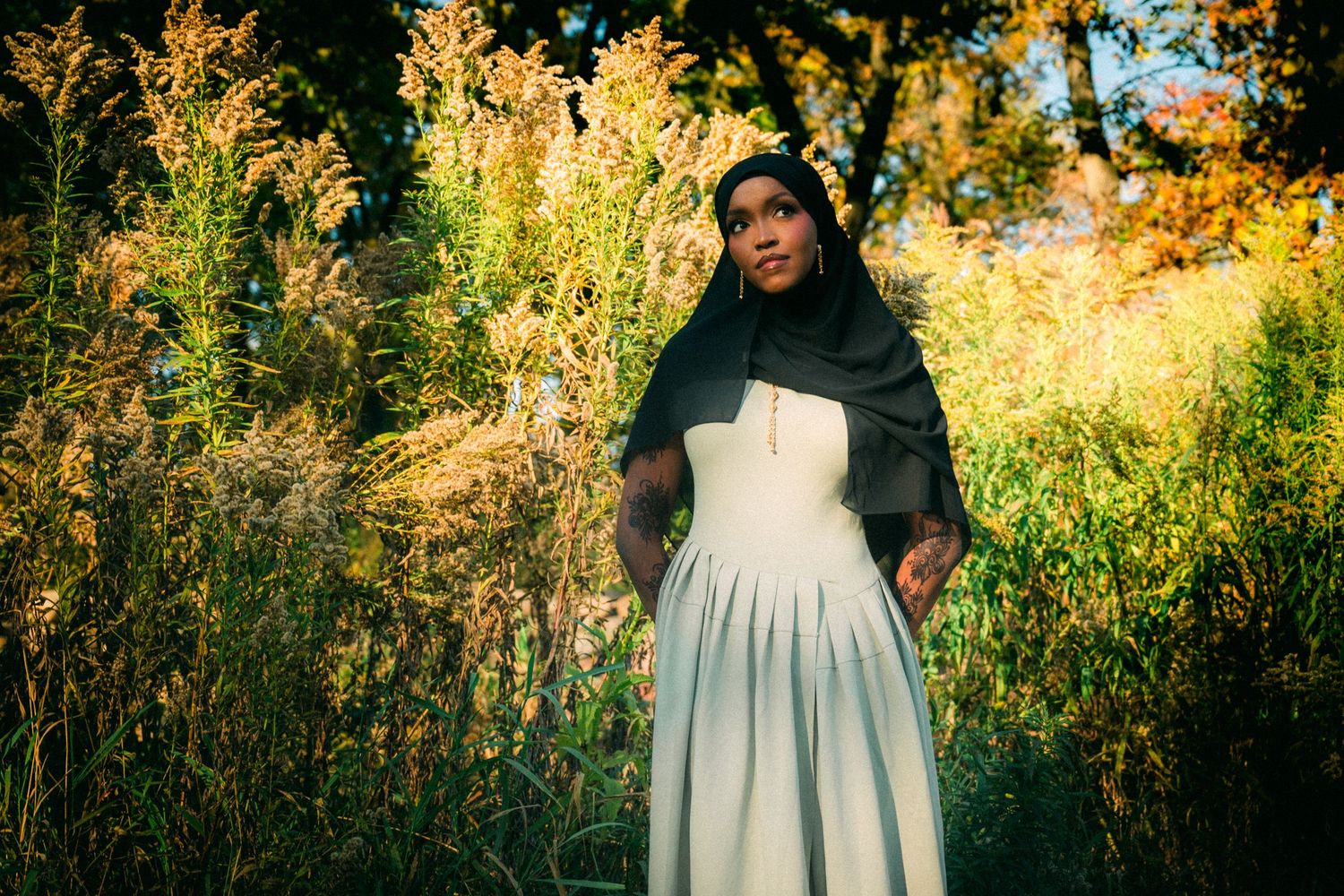 Person in white dress and dark shawl standing among golden wildflowers at sunset.