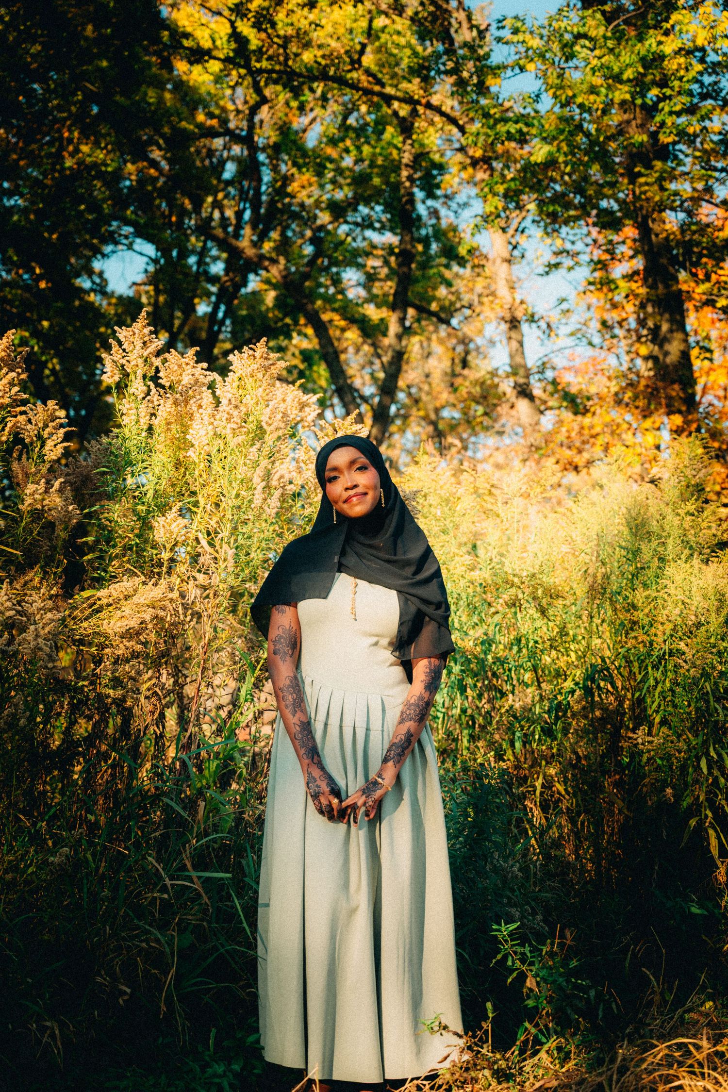 A woman in a hijab and long dress poses outdoors among golden autumn foliage and tall grass.