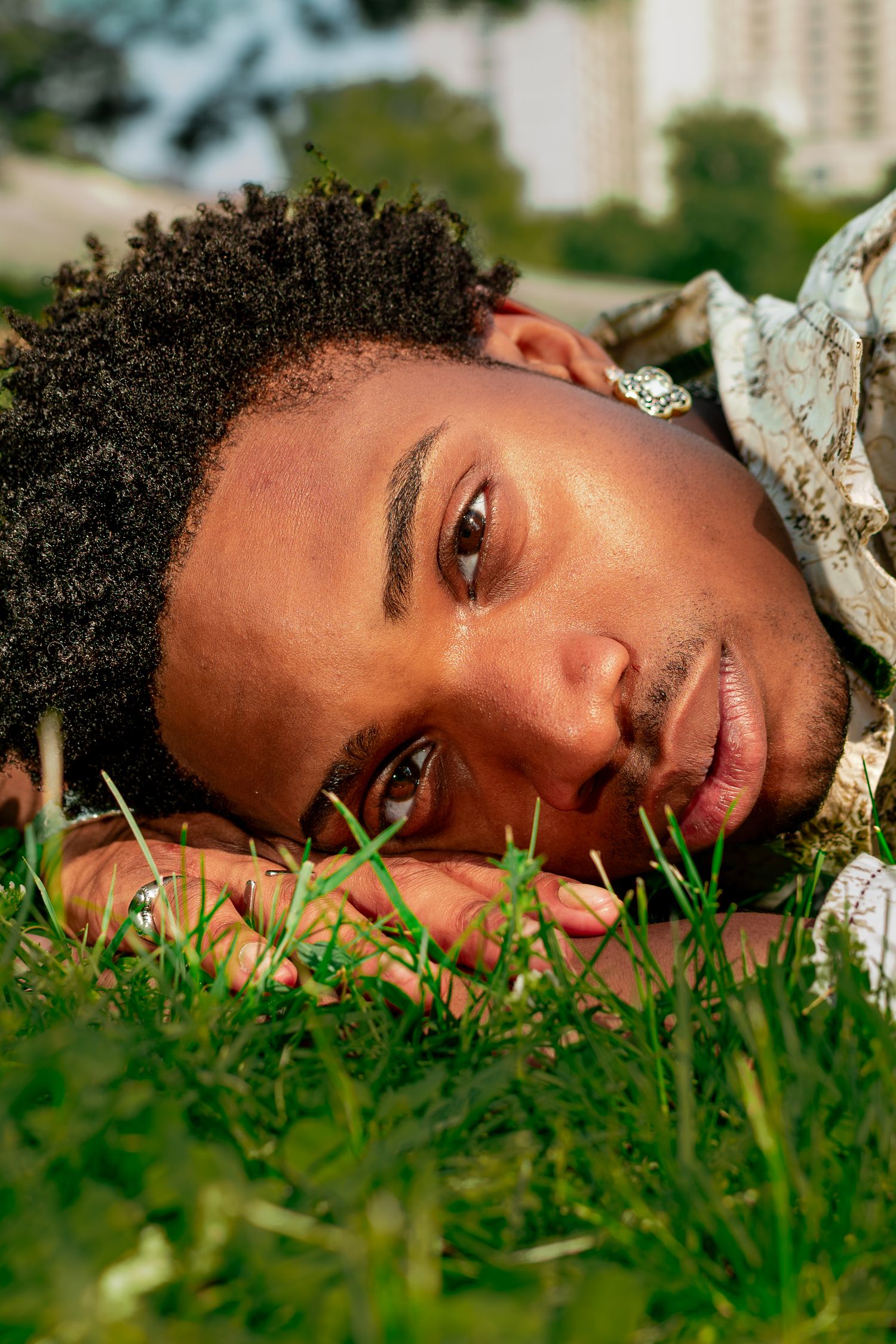 Young Black man with curly hair lying on green grass, gazing at camera with a calm expression outdoors.