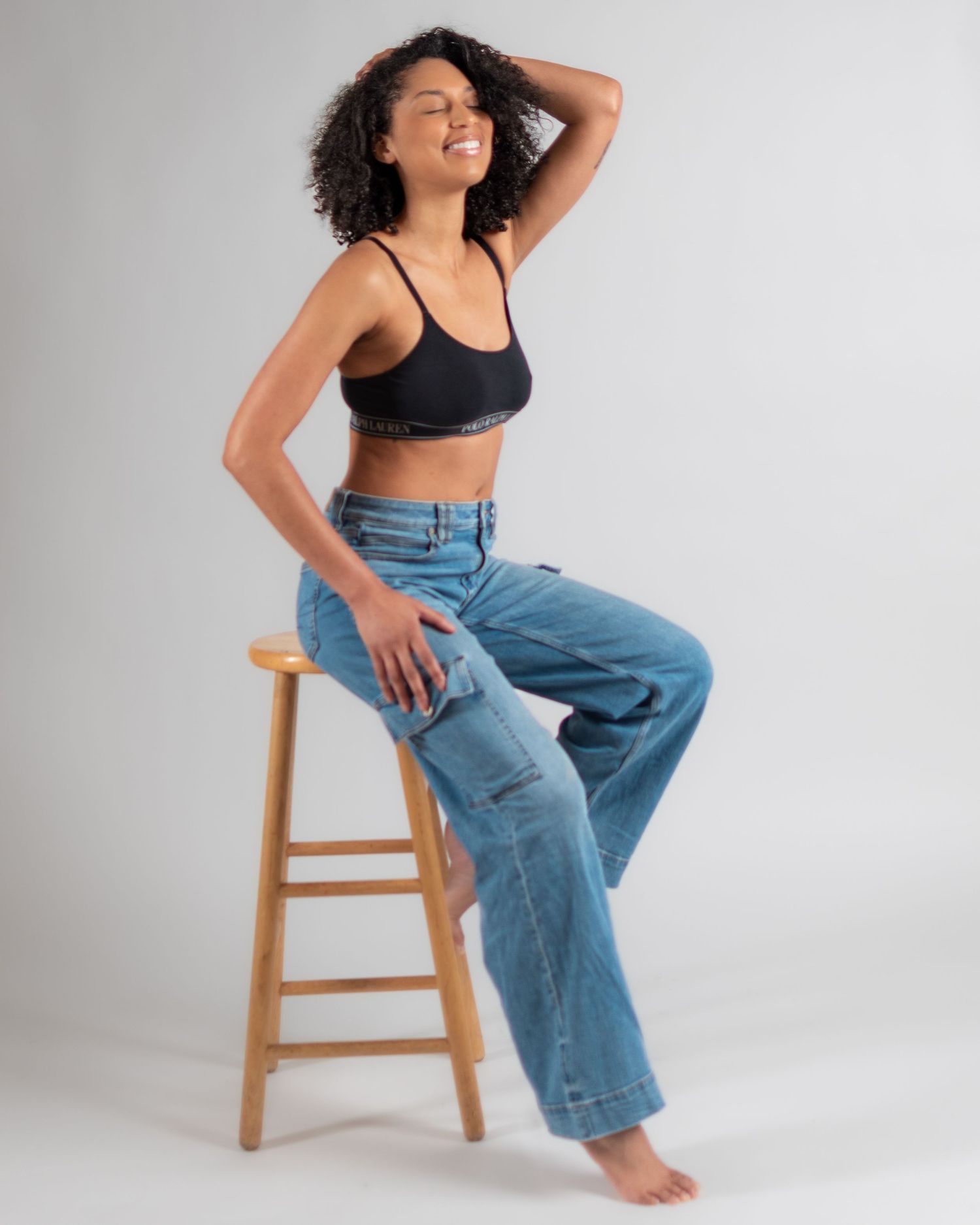 Woman in black crop top and light wash wide-leg jeans sitting on wooden stool, smiling with hand in hair.