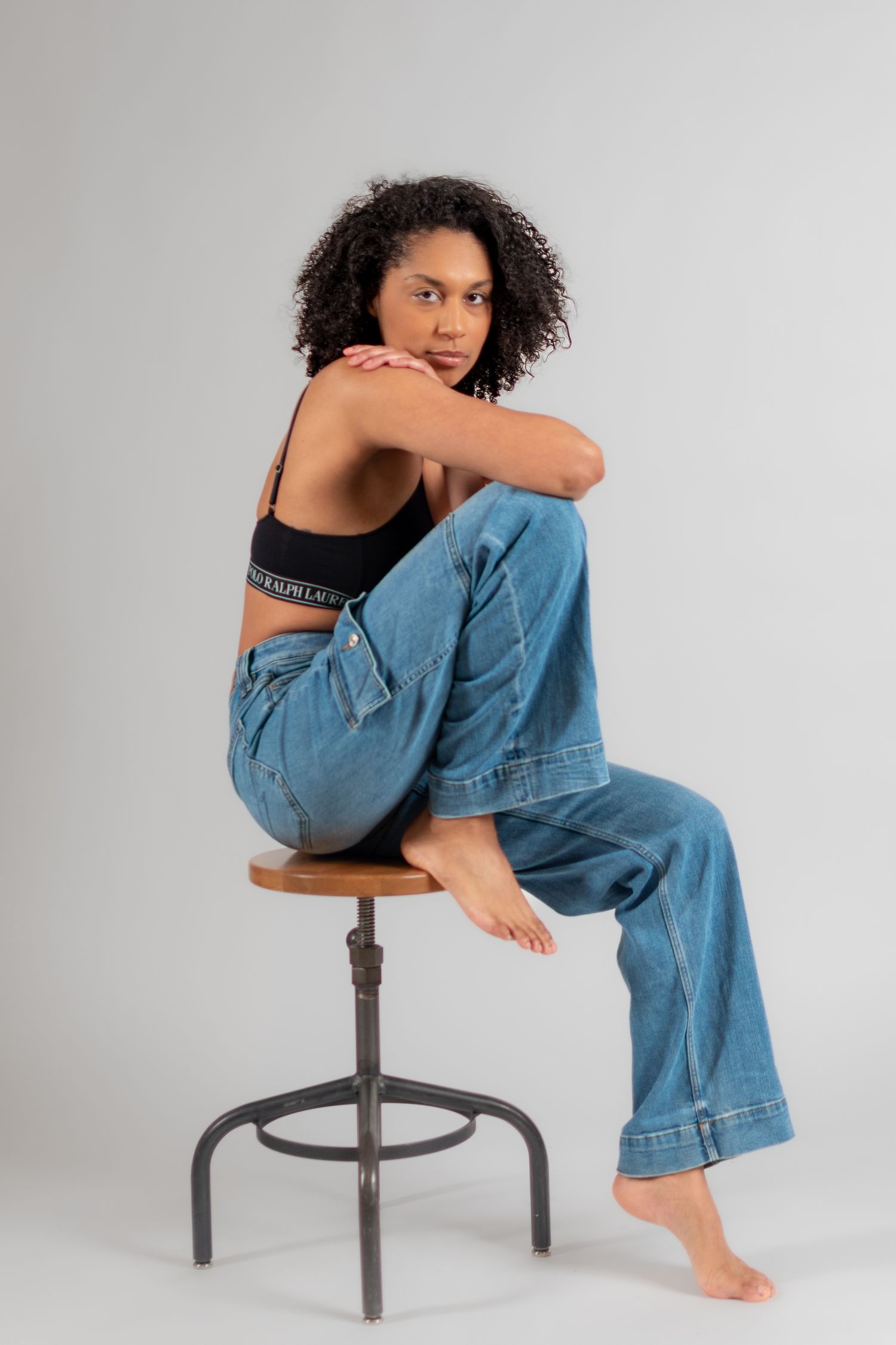 Young woman with curly hair sitting on a stool, wearing a black crop top and wide-leg blue jeans, barefoot.