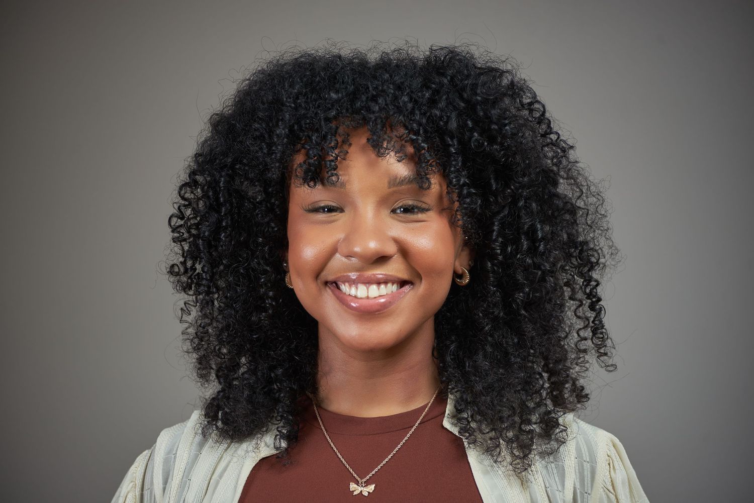 Natural light portrait with curly hair and delicate necklace against grey background.